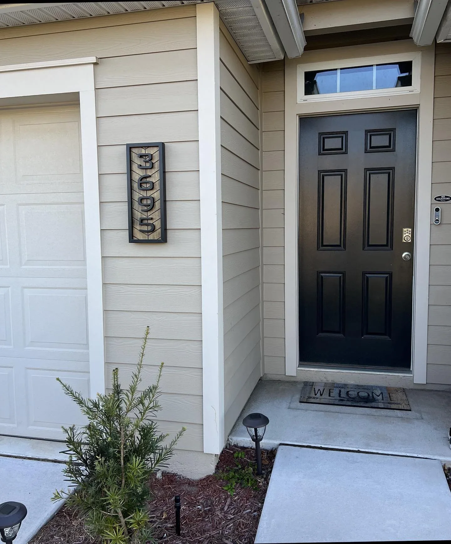 Front porch of a house with a black door, welcome mat, small garden bed, and a sign displaying the house number 1308.