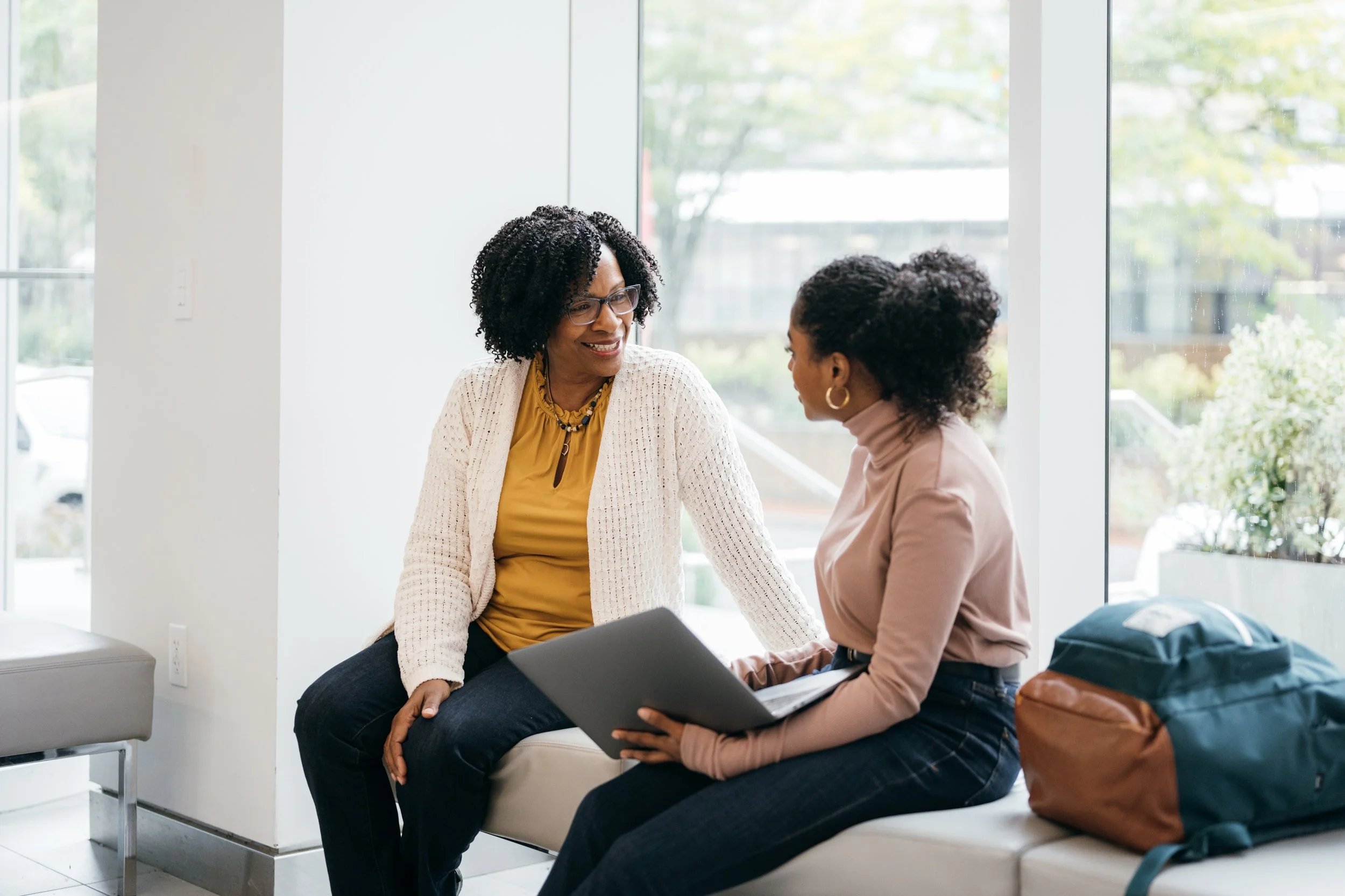 Two women engaging in a conversation in an office with large windows and natural light.