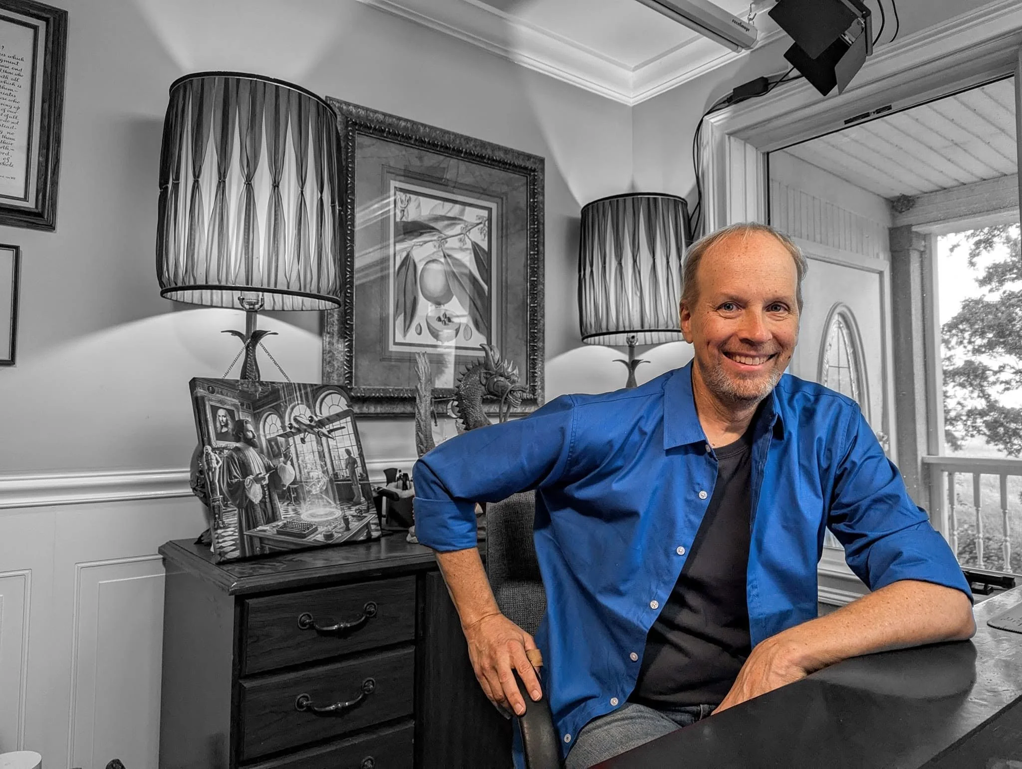 Barclay R. Brown wearing a blue shirt, sitting at a desk in a well-decorated room. The room has framed pictures, a lamp, and a window showing trees outside.