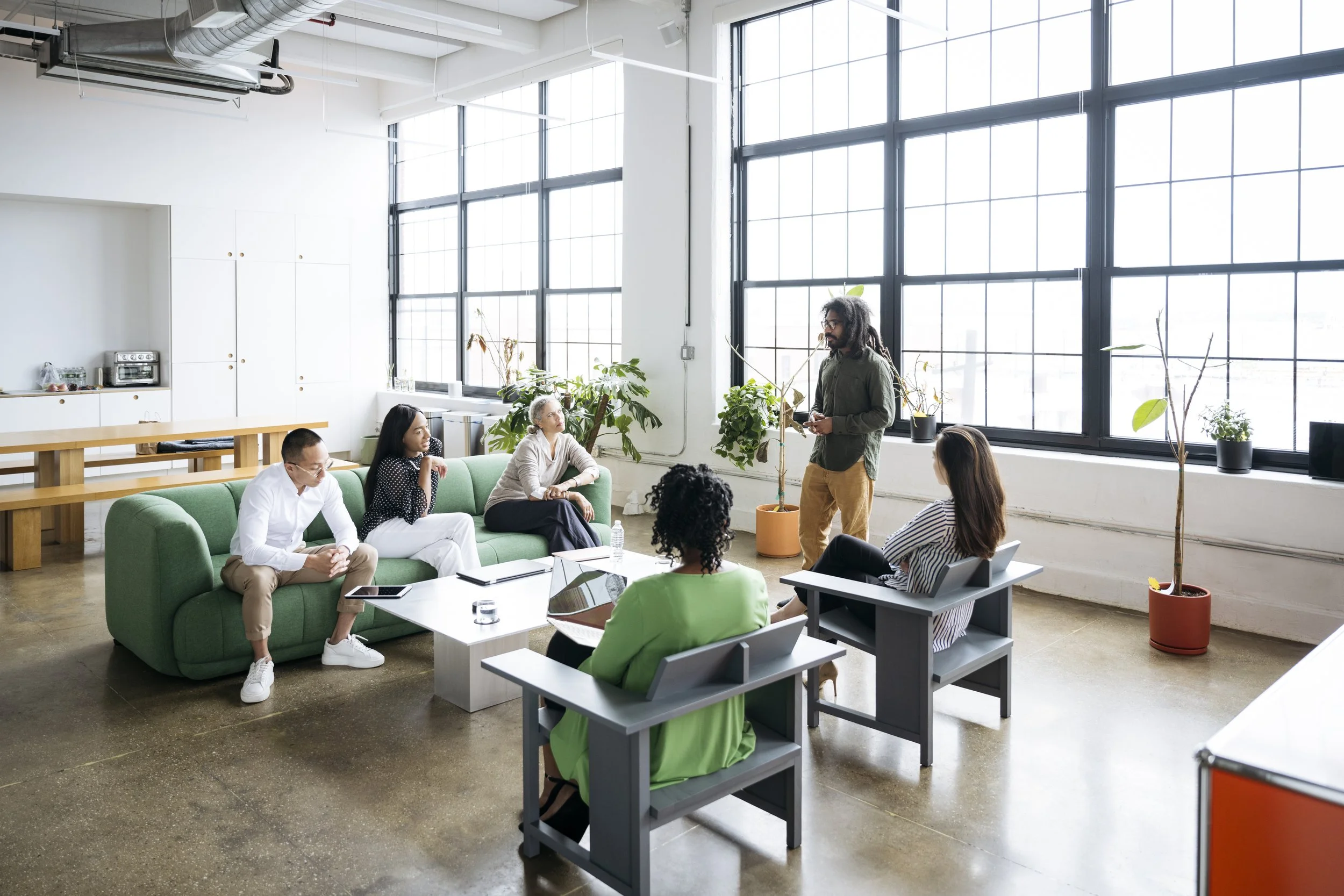 A diverse group of people in a modern, bright office space participate in a discussion or presentation. Some are seated on a green couch and chairs, while a man with dreadlocks, wearing a green shirt and khaki pants, stands presenting. The office has large windows, potted plants, and minimalist decor.