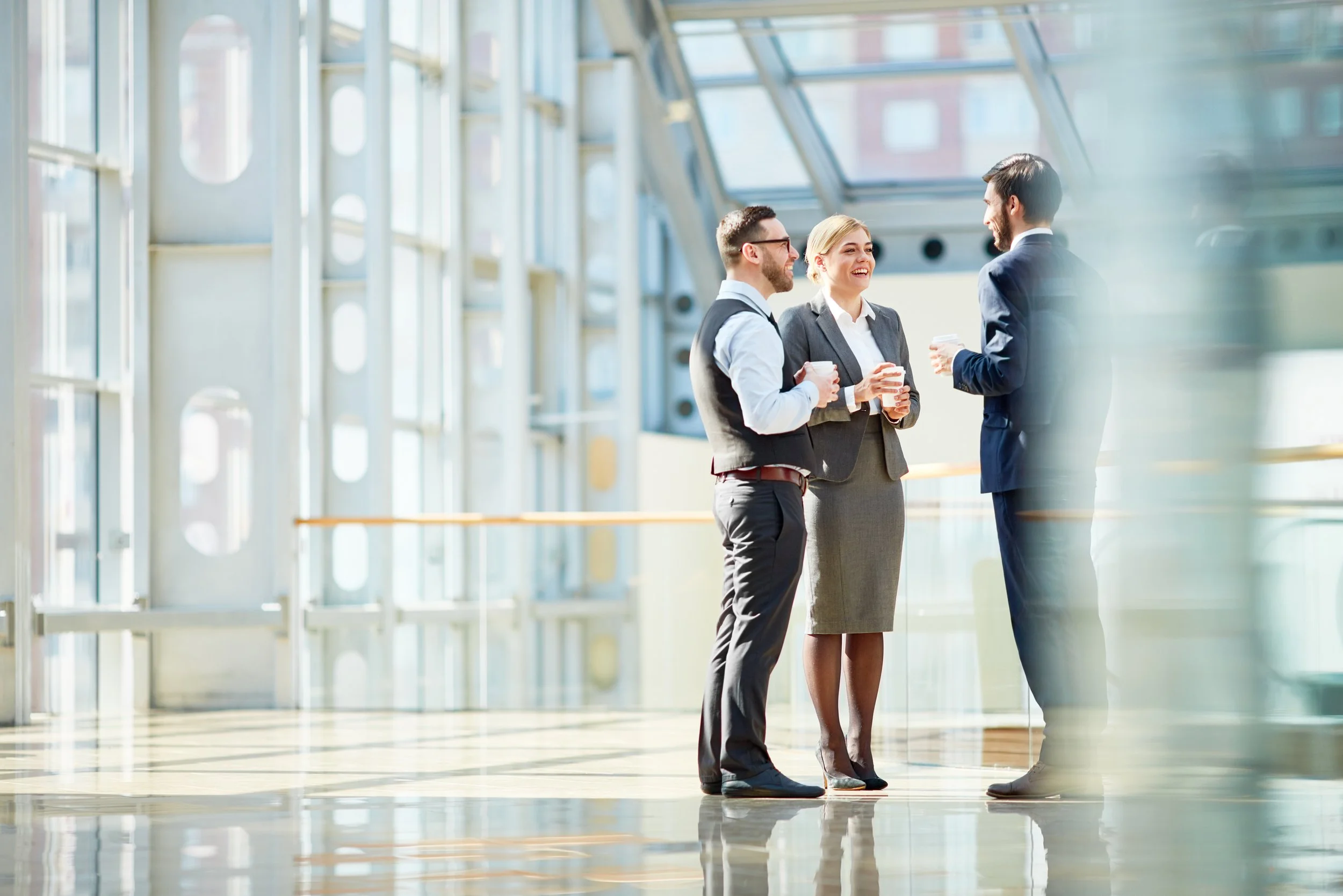 Three business professionals, two men and one woman, are standing and chatting inside a modern building with large glass windows. They appear to be having a conversation while holding coffee cups.