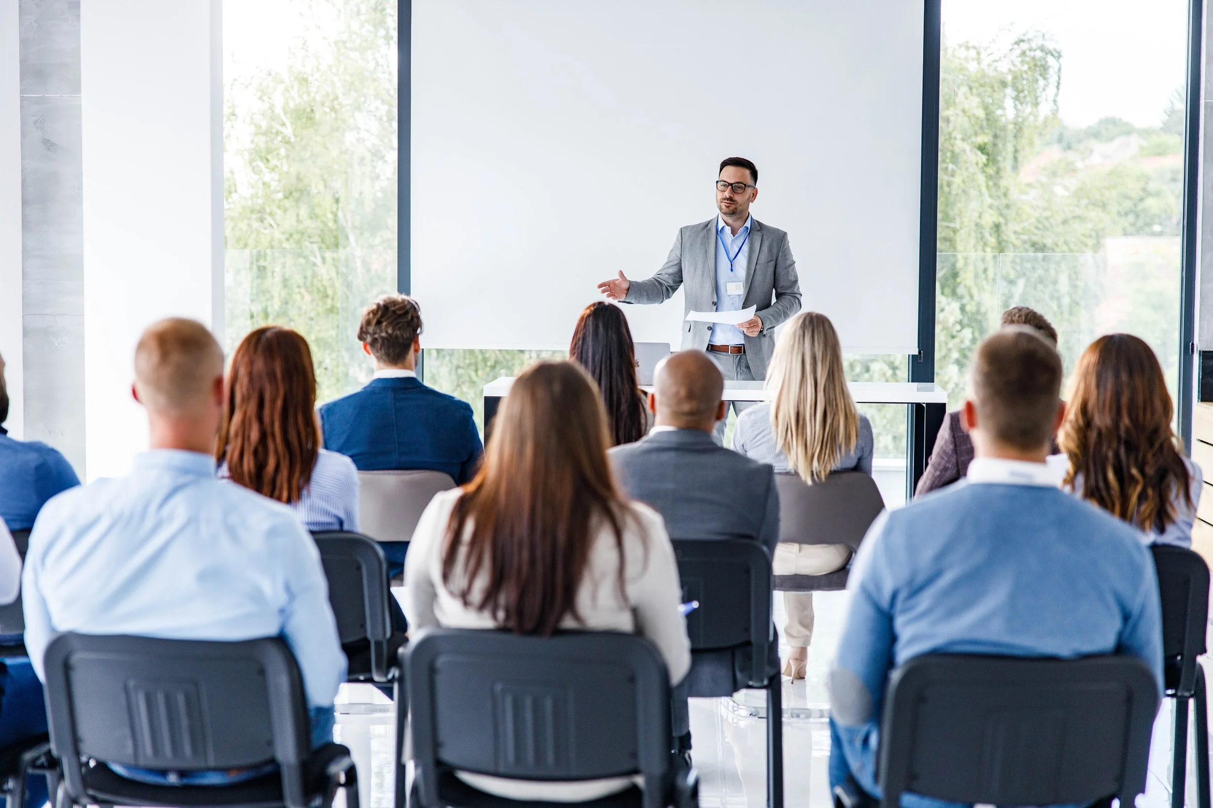 Man giving a presentation to an audience in a bright room with large windows and a blank white screen.