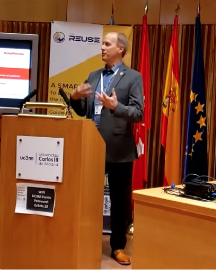 Barclay R. Brown speaking at a podium during a presentation, with flags of Spain, European Union, and other countries behind him.