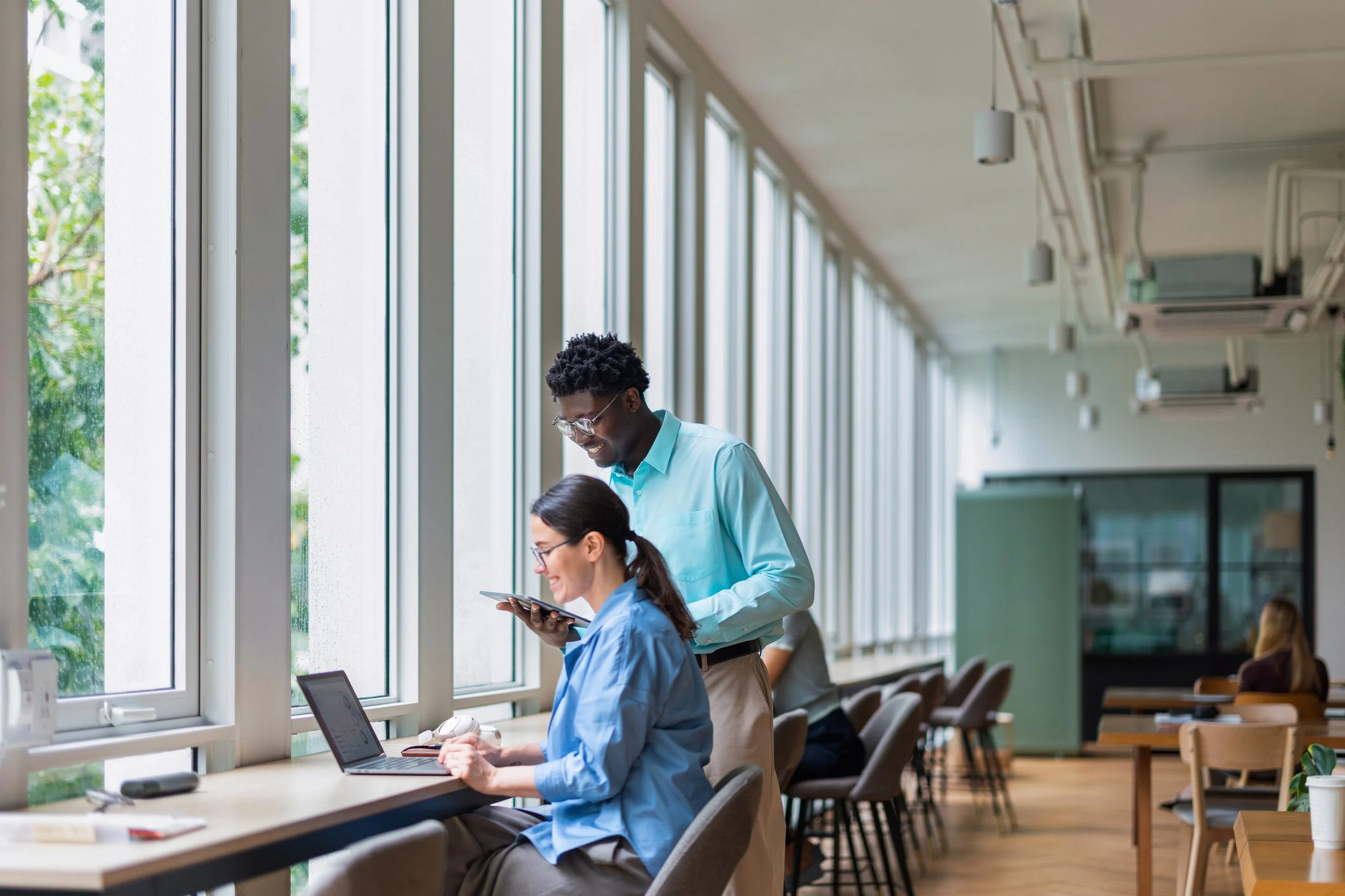 People working on laptops and tablets inside a bright, modern classroom or library with large windows.