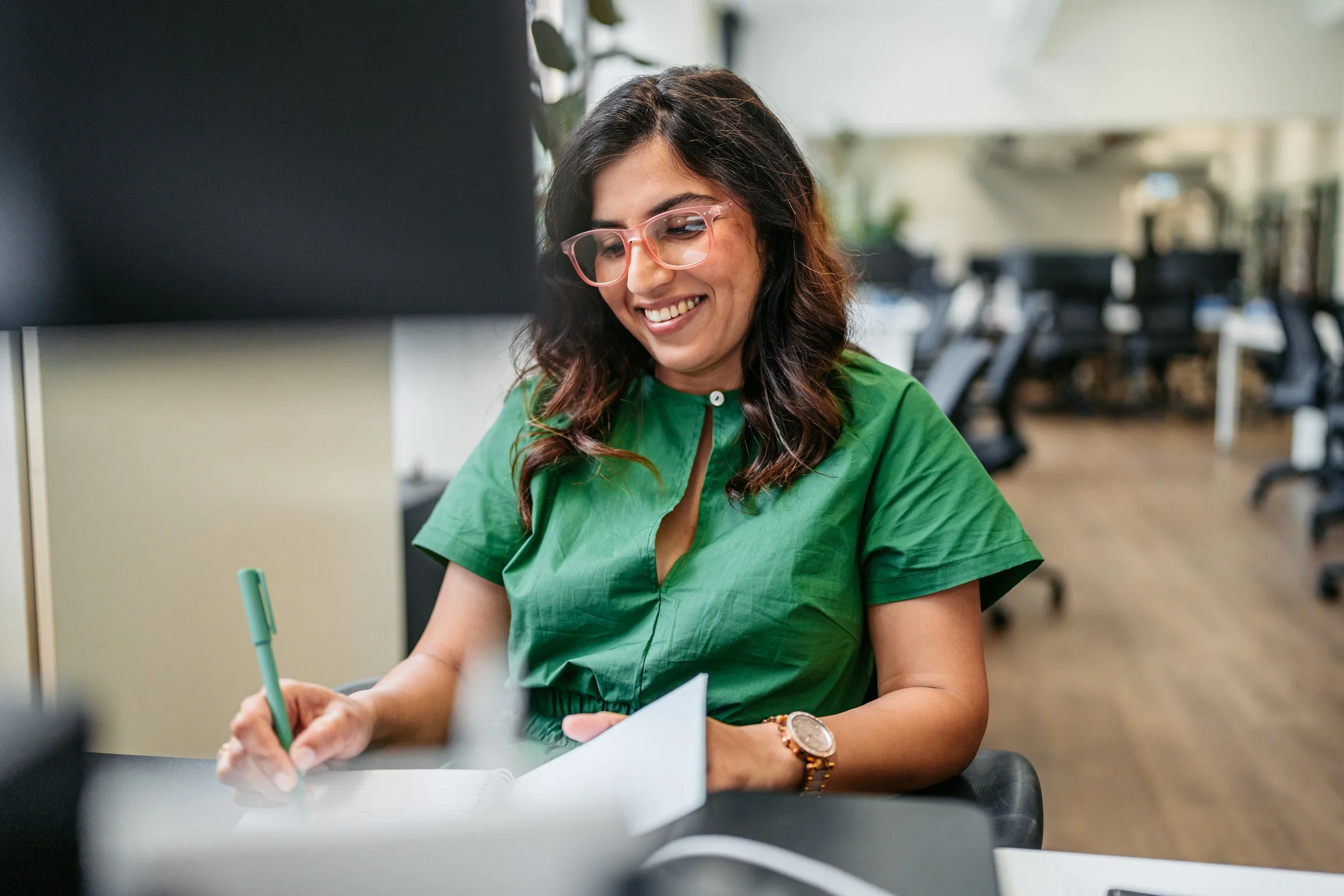 A woman with dark, wavy hair wearing glasses and a green top, smiling while writing on a notepad at a desk in an office environment.