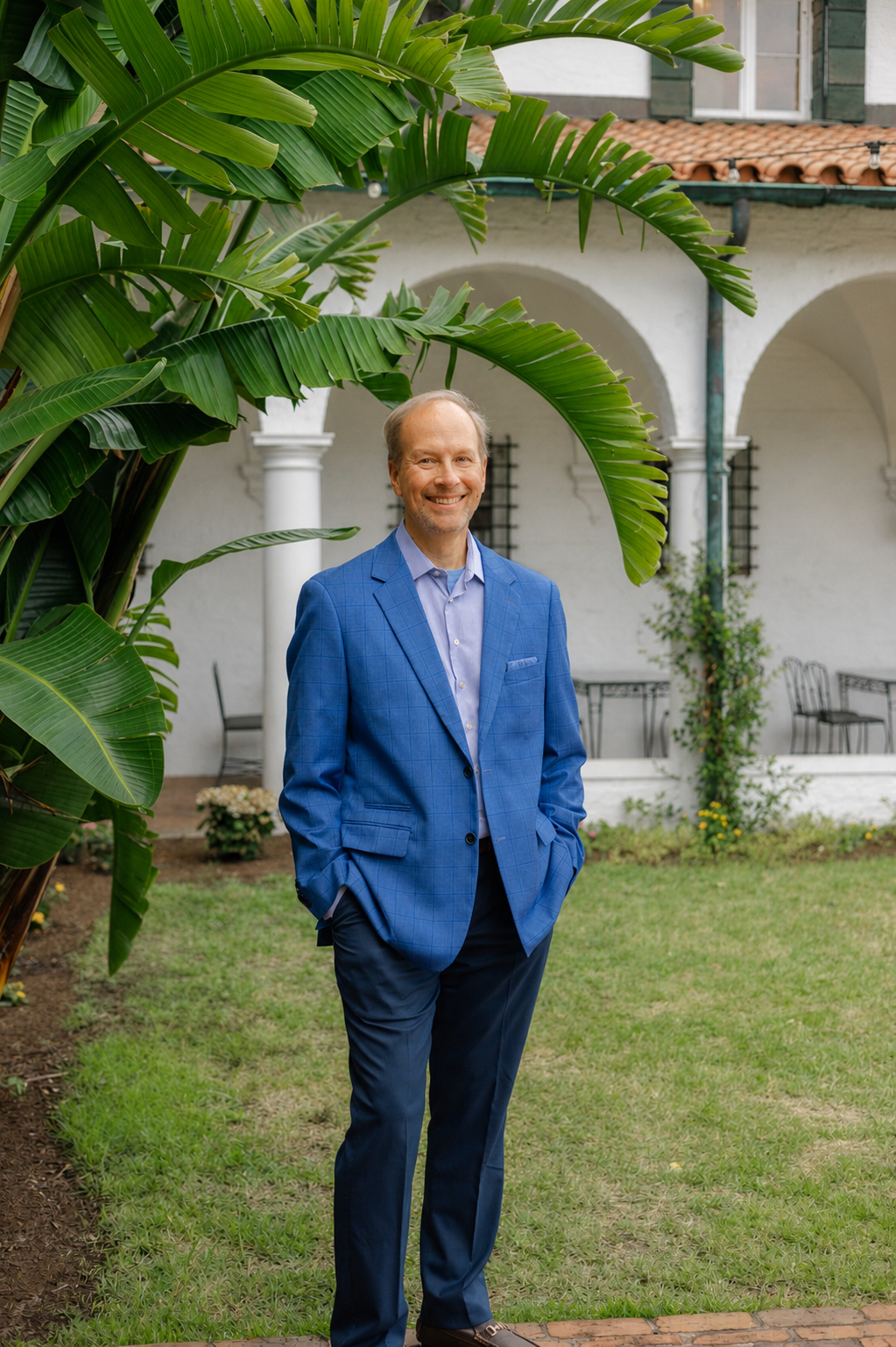 Barclay R. Brown standing outdoors in front of a white building with arches. Large green tropical leaves partially frame his face and upper body.