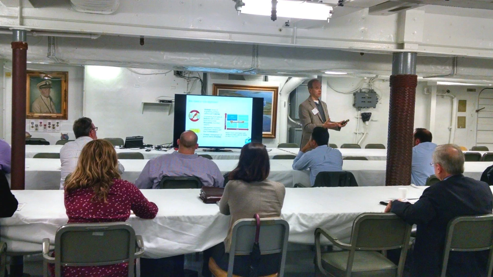 A man giving a presentation to a group of people seated around conference tables in a basement or industrial space, with a large screen displaying a slide, portraits on the wall, and exposed pipes and electrical panels.