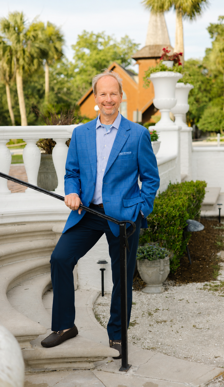 A man in a blue suit standing on outdoor stairs with a white ornate balustrade, smiling, with a house and trees in the background.