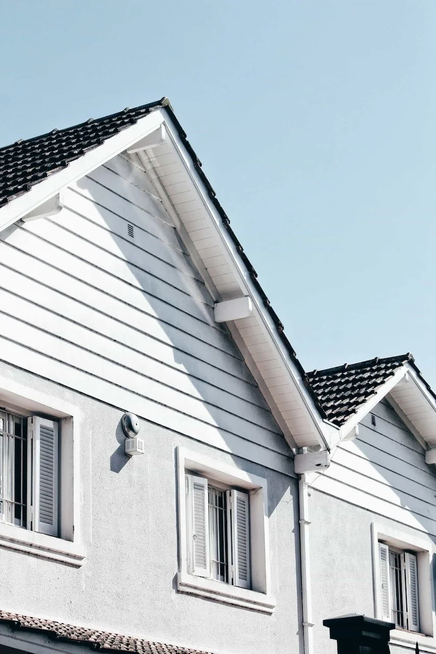 Close-up of the upper part of a house with a white exterior, showing windows with shutters, a small vent, and a wall-mounted outdoor light fixture, under a clear blue sky.