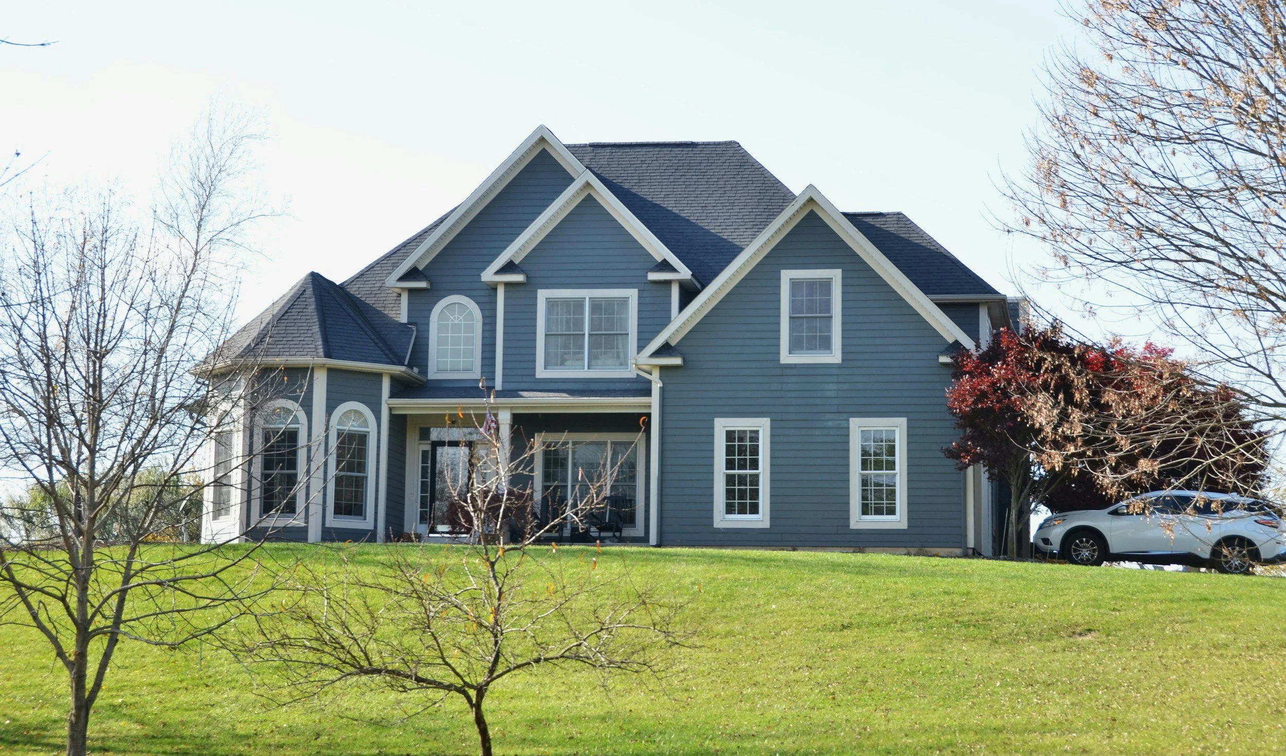 A large blue house with multiple gabled roofs and white trim, surrounded by trees with some leaves fallen, and a green lawn in the foreground. A car is parked on the driveway to the right.