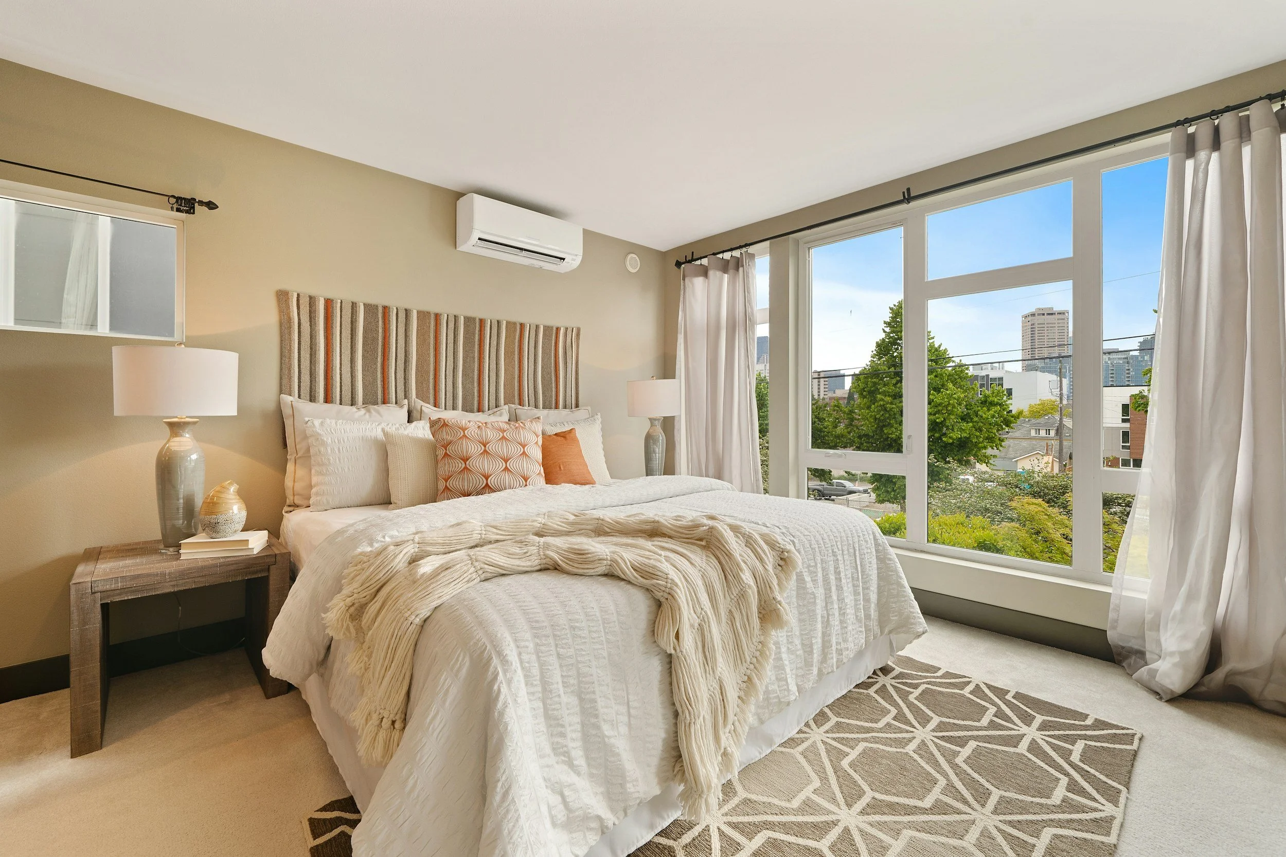 A bedroom with a large window showing a cityscape and trees outside. The bed has a white quilt, multiple pillows, and a beige throw blanket. There are two nightstands with lamps beside the bed. The room has beige walls, white curtains, and a patterned rug on the floor. An air conditioning unit is mounted on the wall.