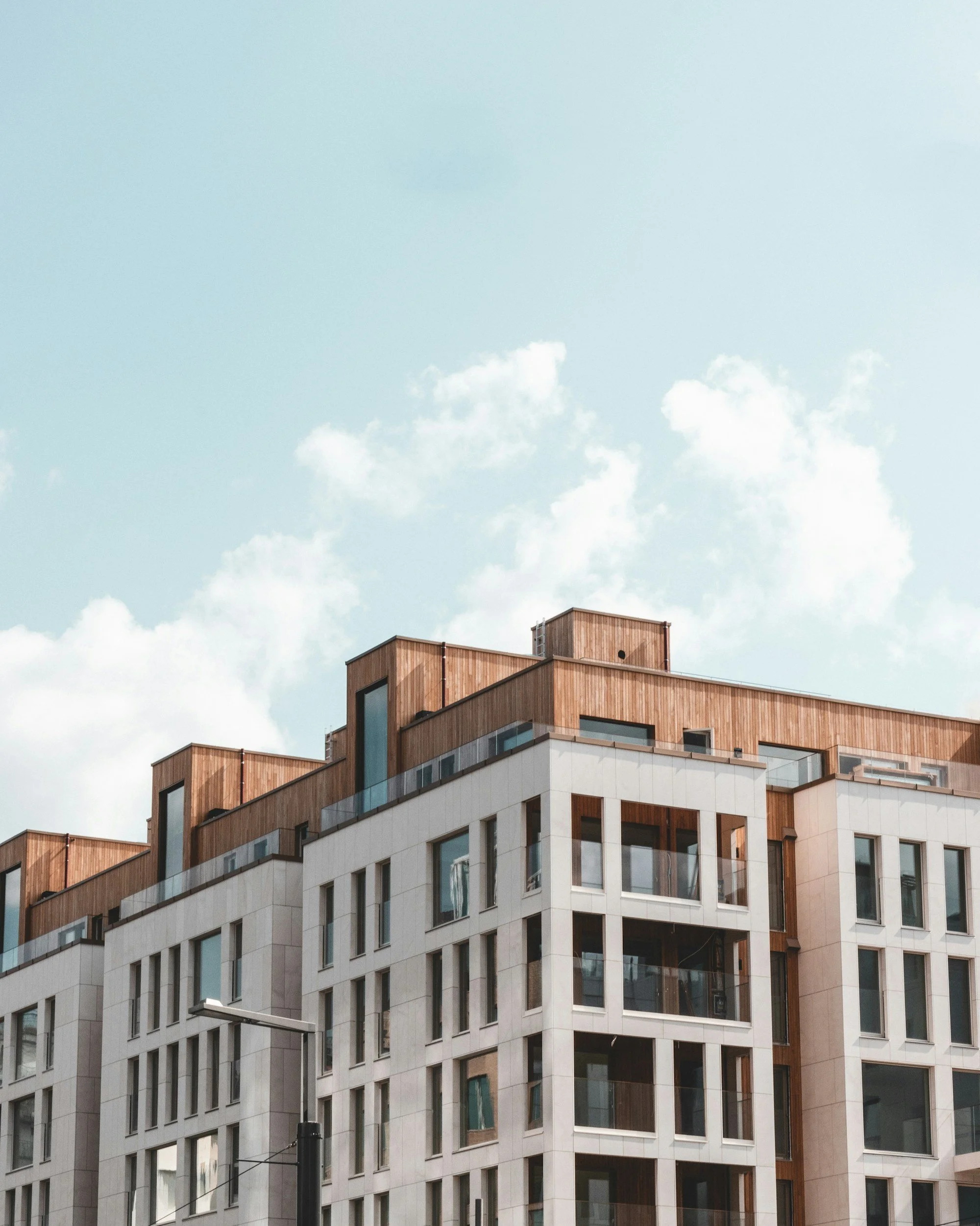 A multi-story modern building with a mix of white and wood exterior, large windows, and a clear blue sky with some clouds above.