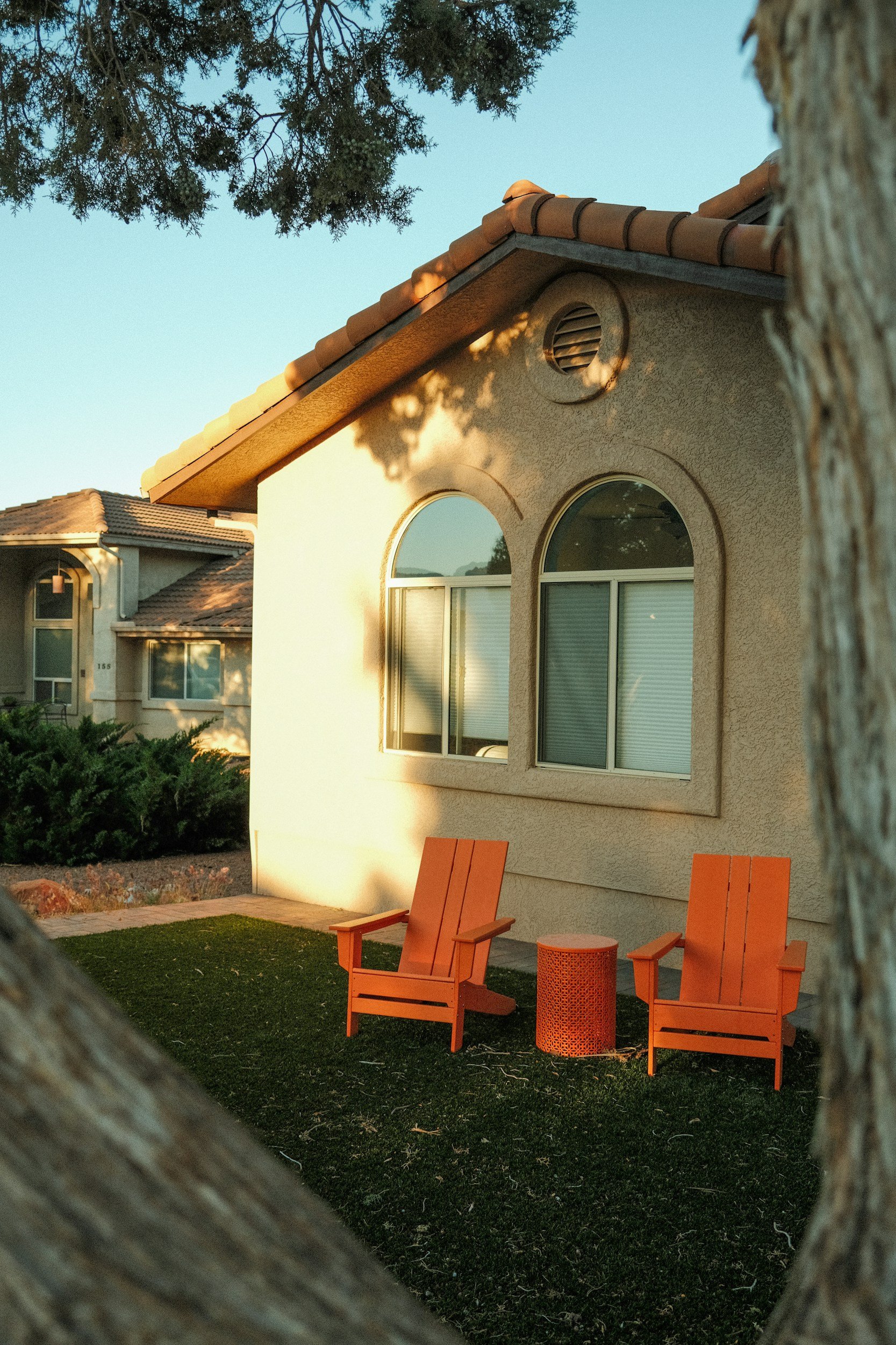 View of a house with beige walls, arched windows, and a tiled roof, seen through a frame of tree branches and a wooden fence. There are three orange chairs and an orange table on a small lawn in the backyard, with bushes and a paved pathway.