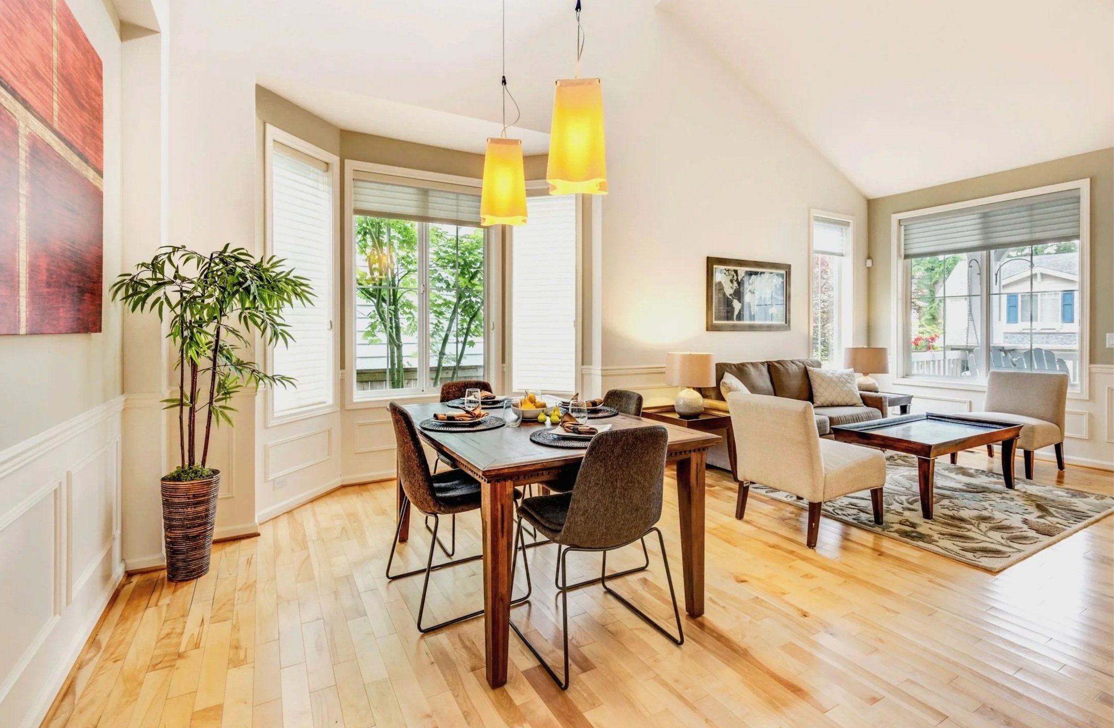 A bright living and dining room with hardwood floors, large windows with white blinds, a wooden dining table set for four, a beige sofa, and a white armchair. There are three yellow pendant lights hanging from the ceiling, a tall potted plant in the corner, and artwork on the walls. Outside the windows, greenery and neighboring houses are visible.