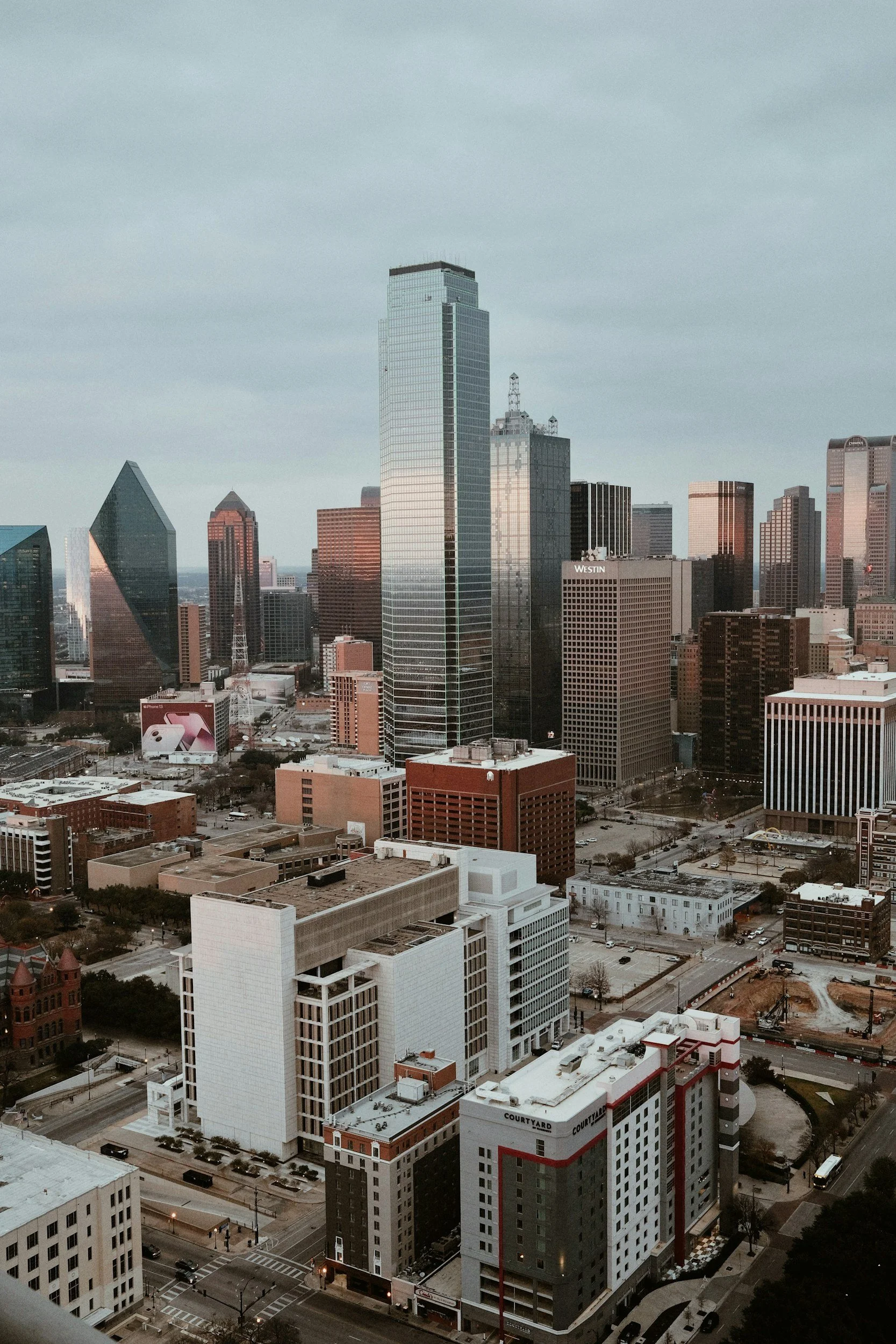 A city skyline with multiple modern skyscrapers under a cloudy sky, including tall glass buildings and mid-rise structures, with streets and parking lots in the foreground.