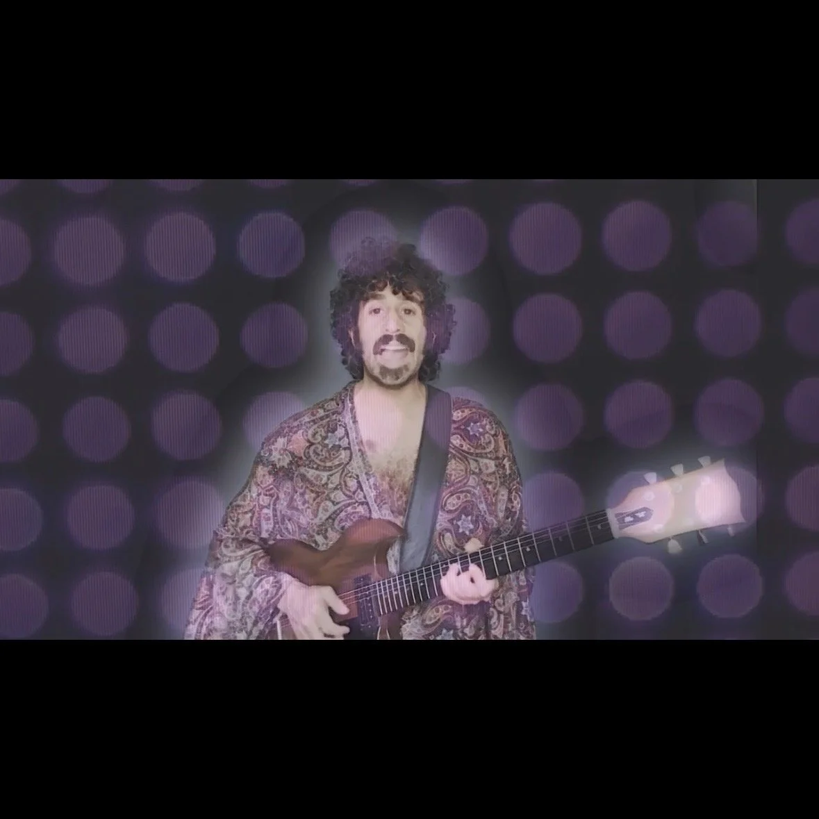 A man with curly hair and a beard holding a guitar, standing in front of a background with purple circular lights.