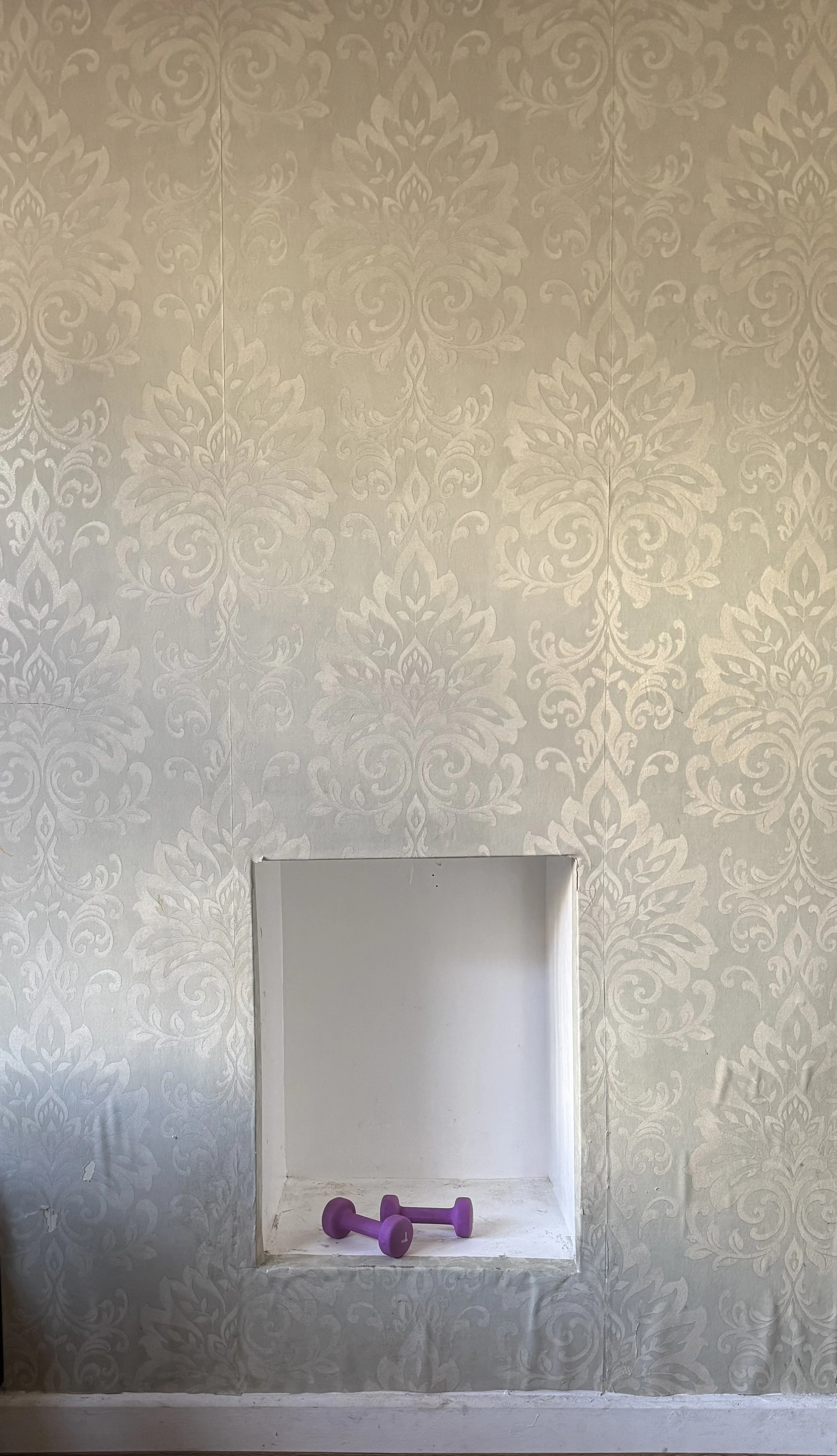 A white wall with ornate, patterned wallpaper surrounding a small rectangular niche or window opening. In front of the opening, there are two small purple dumbbells on the floor.