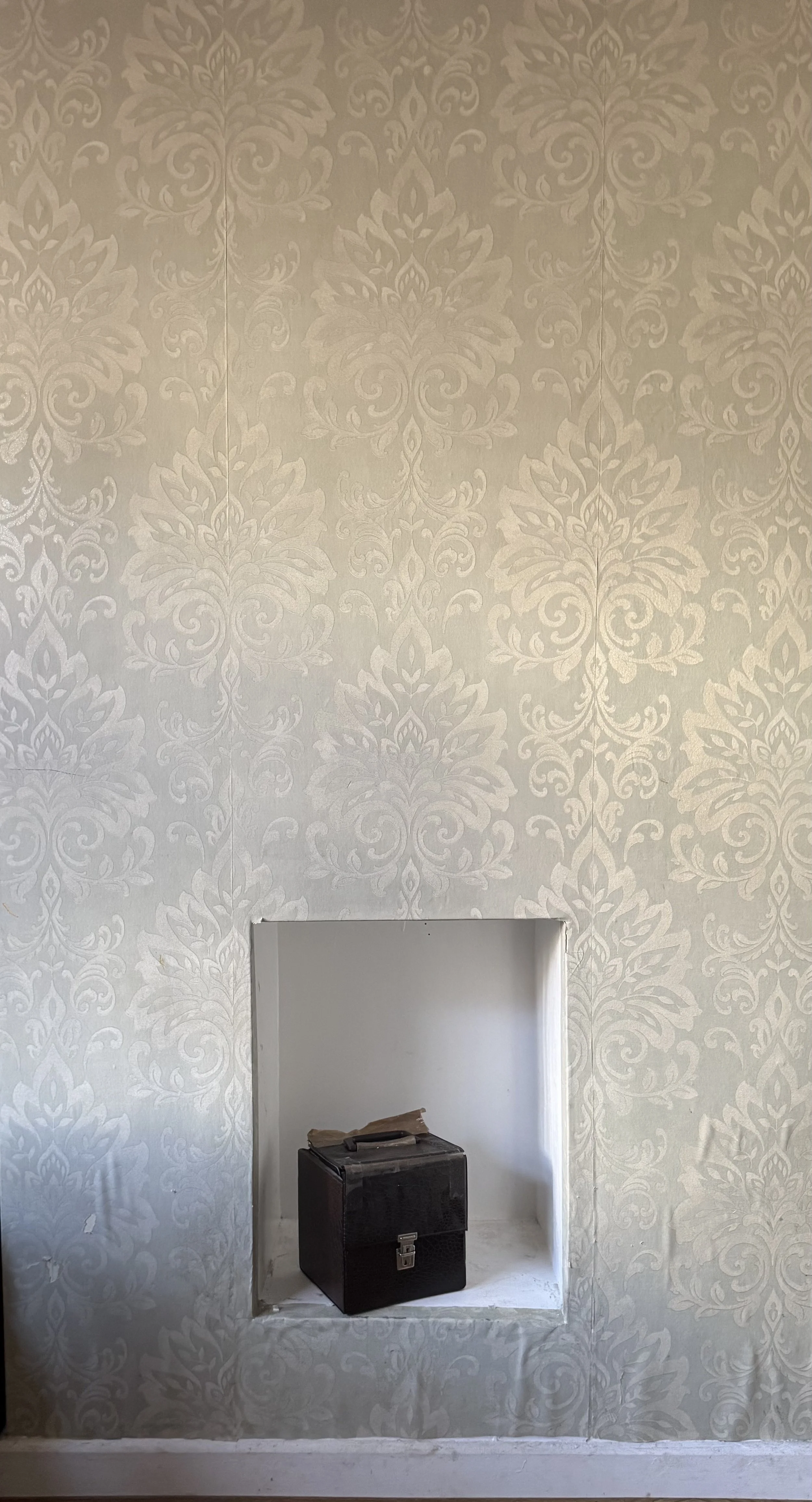 A room with ornate beige wallpaper with a floral pattern. There is a small recessed niche in the wall with a black box or speaker and some wood on top inside the niche.
