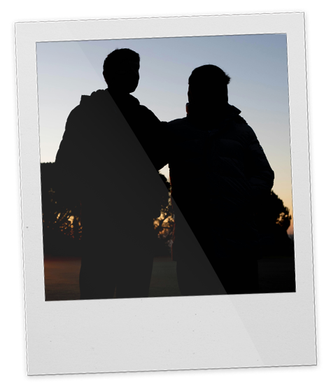 Two people, a father and a son, standing outdoors during sunset, one with an arm around the other, silhouetted against the sky.