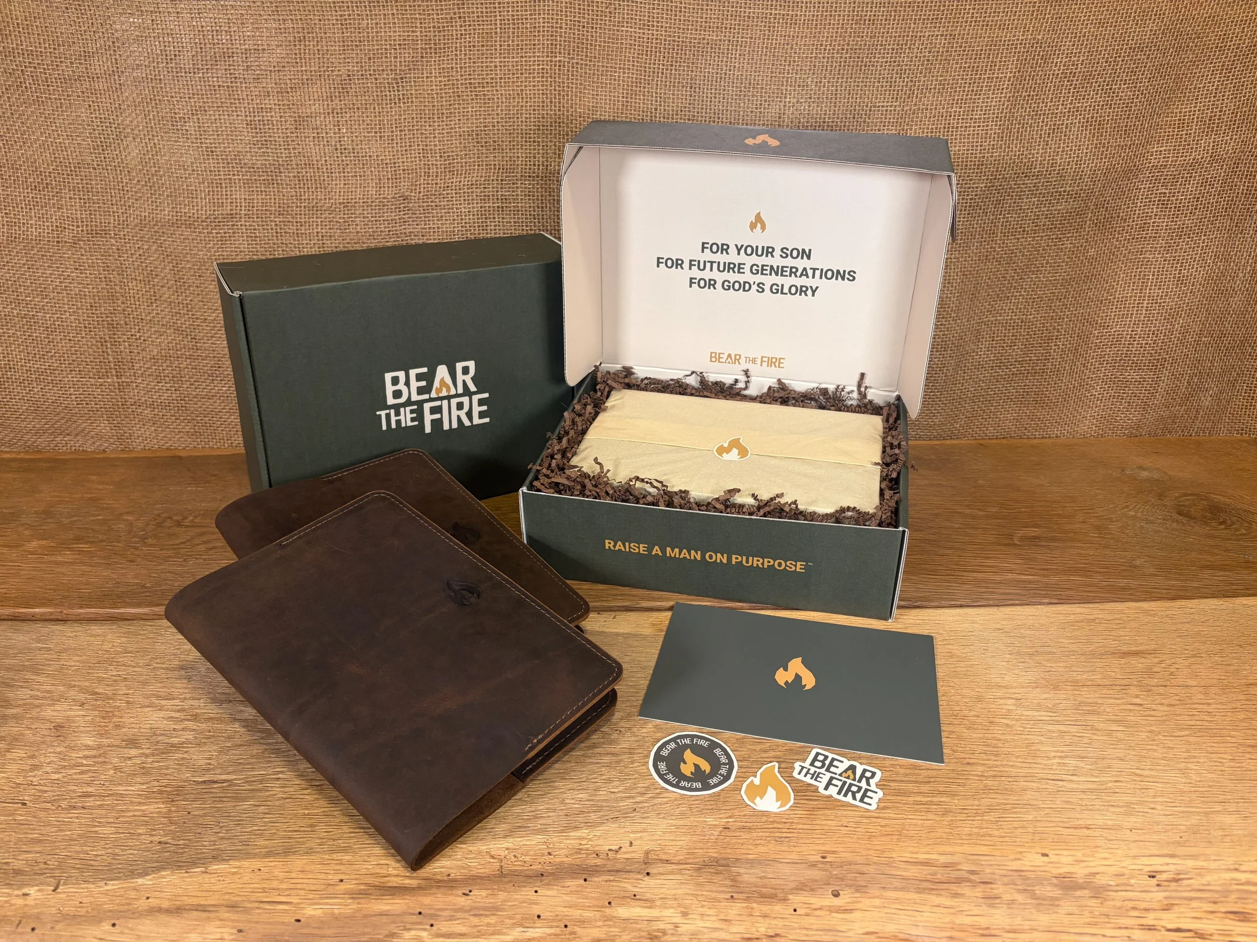 Open gift box with the message "For your son, for future generations, for God's glory" inside, surrounded by stickers and two leather journals on a wooden surface with a burlap background.
