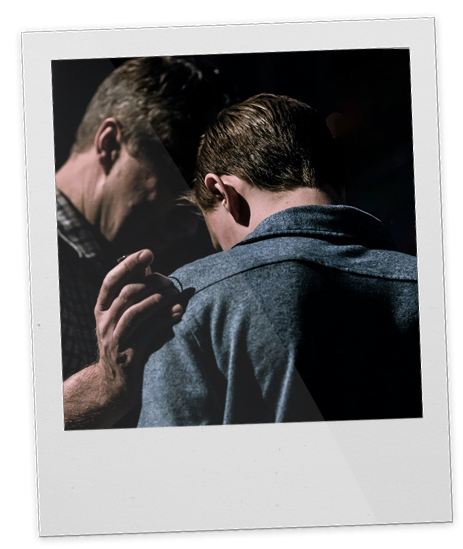 Two men face each other closely, a father and a son, praying together. One appears to be touching the other's shoulder in a comforting or intimate gesture, with a dark background and soft lighting.