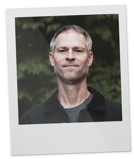 Portrait of a middle-aged man with short gray hair and a slight beard, wearing a dark jacket, standing outdoors with blurred greenery in the background.