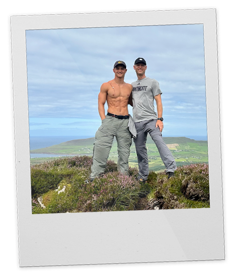 Two men standing together outdoors on a hilltop with a scenic landscape of water and green fields in the background.
