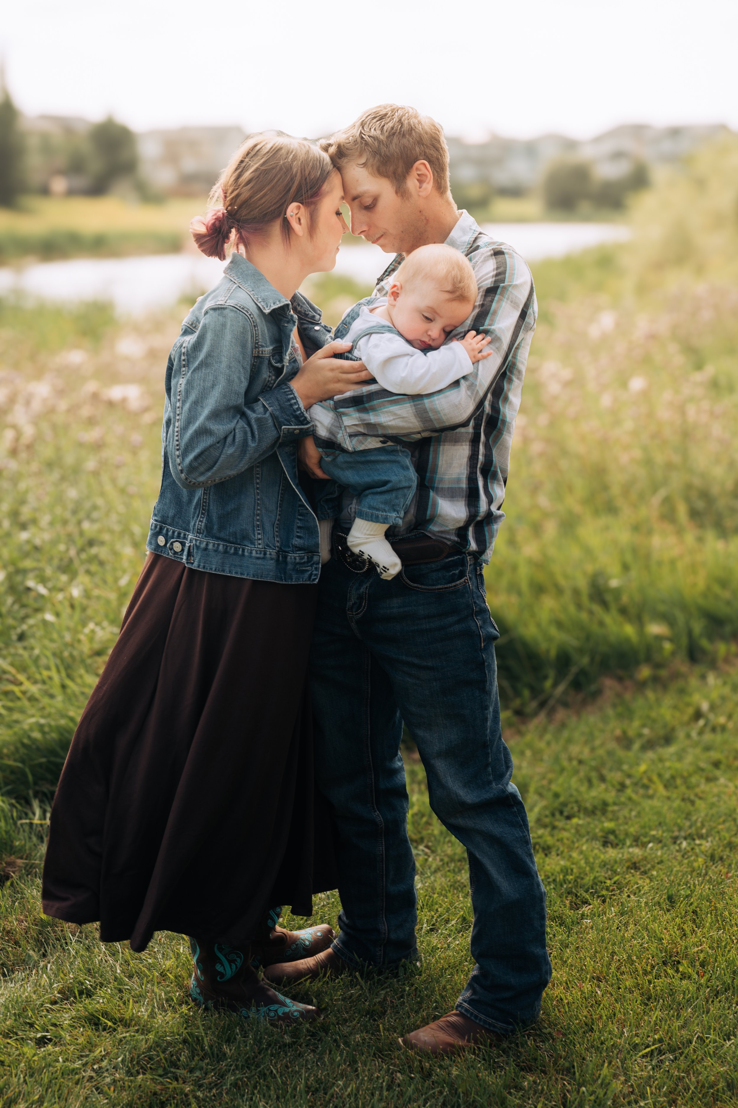 A young couple and a baby standing outdoors in a grassy area near a river, with their heads touching and eyes closed, creating an intimate and tender moment.