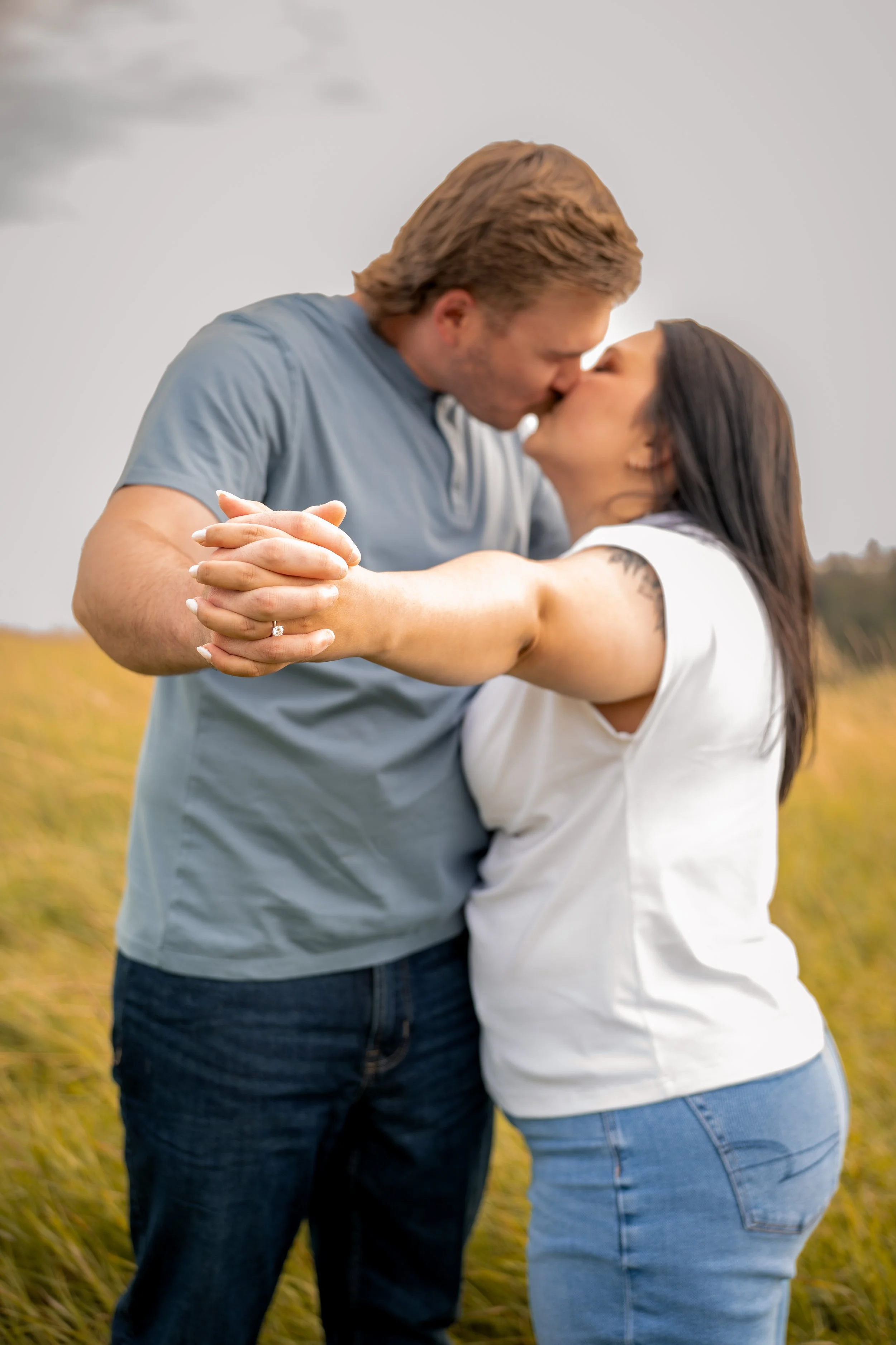 A couple kissing outdoors in a grassy field, with the woman wearing a white shirt and blue jeans, and the man wearing a gray shirt and dark jeans.