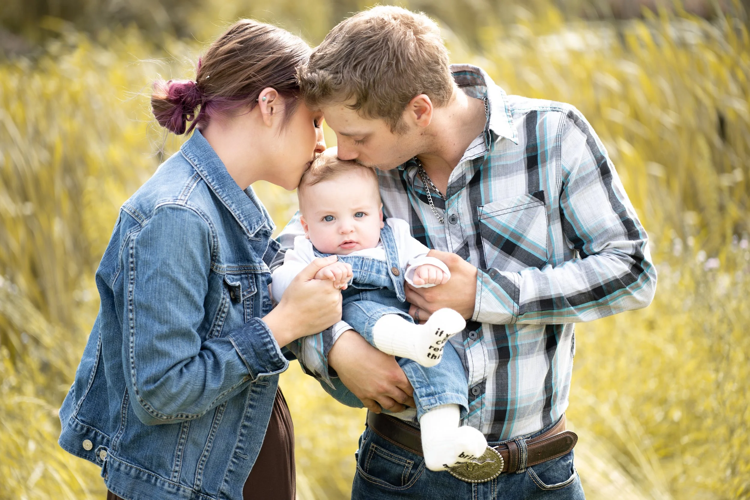 A young woman and a young man are holding a baby together outdoors, with their foreheads touching and eyes closed, as if sharing a tender moment. The woman has purple-tinted hair styles in a bun, and the man has light brown hair. They are dressed cas