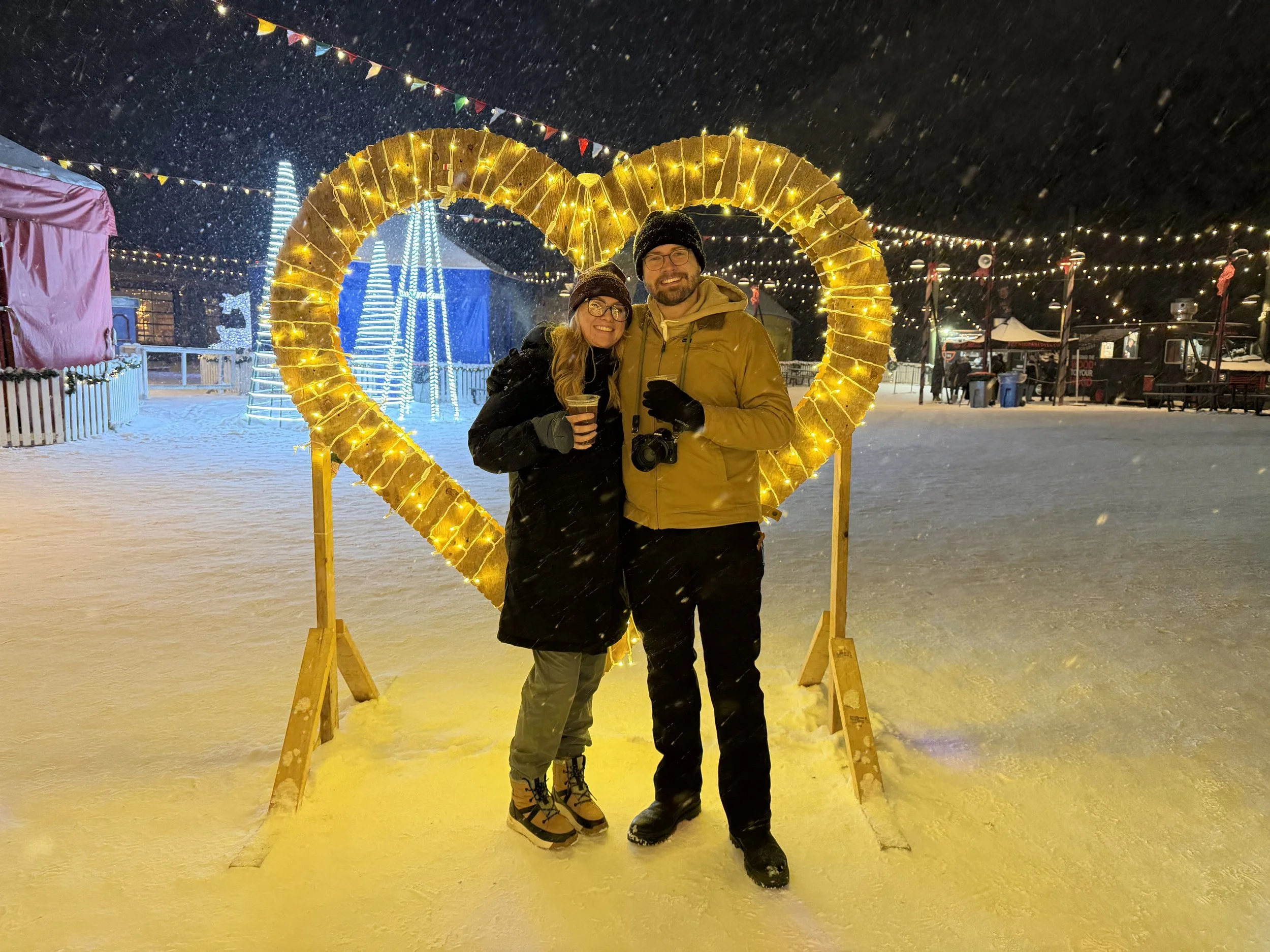 Two people, a woman and a man, smiling and standing together at a festive winter outdoor event, framed by a large illuminated heart in the snow, with decorative lights and tents in the background.