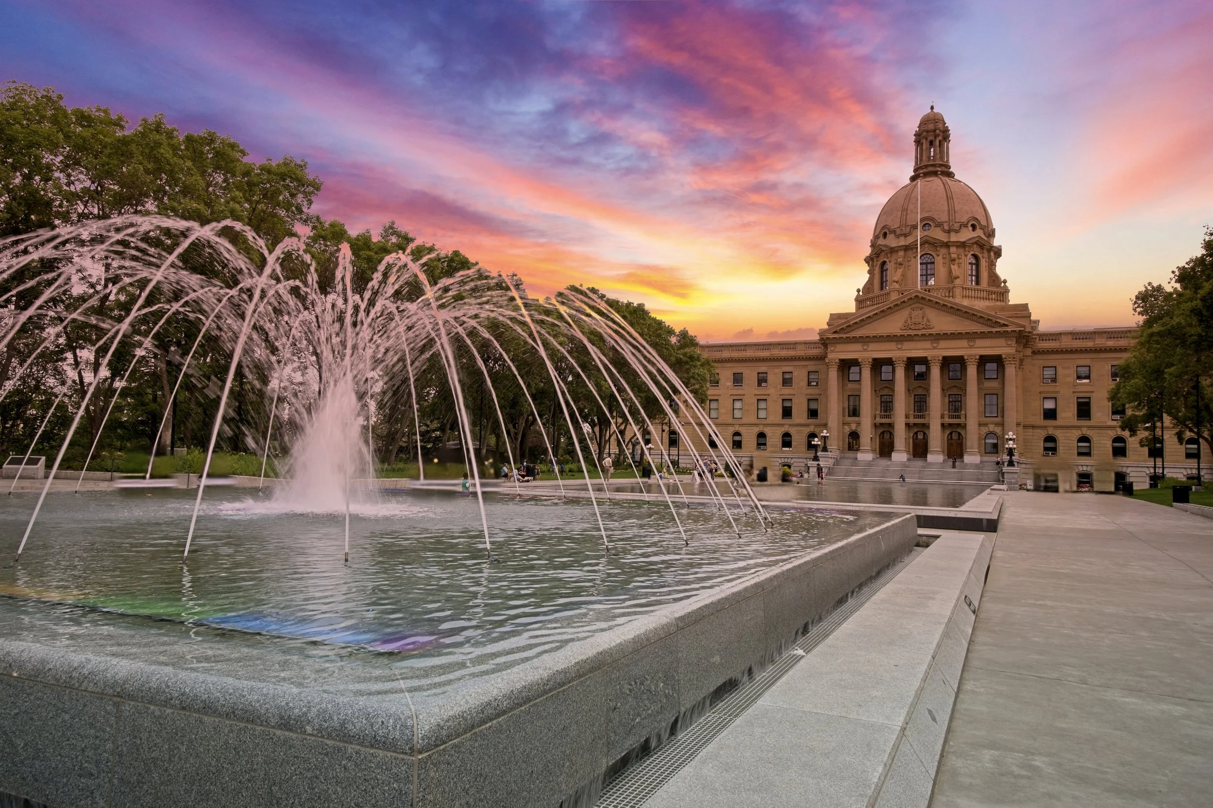 A historic government building with a dome and columns, surrounded by trees, under a colorful sunset sky. In the foreground, a fountain with water jets is part of a plaza.