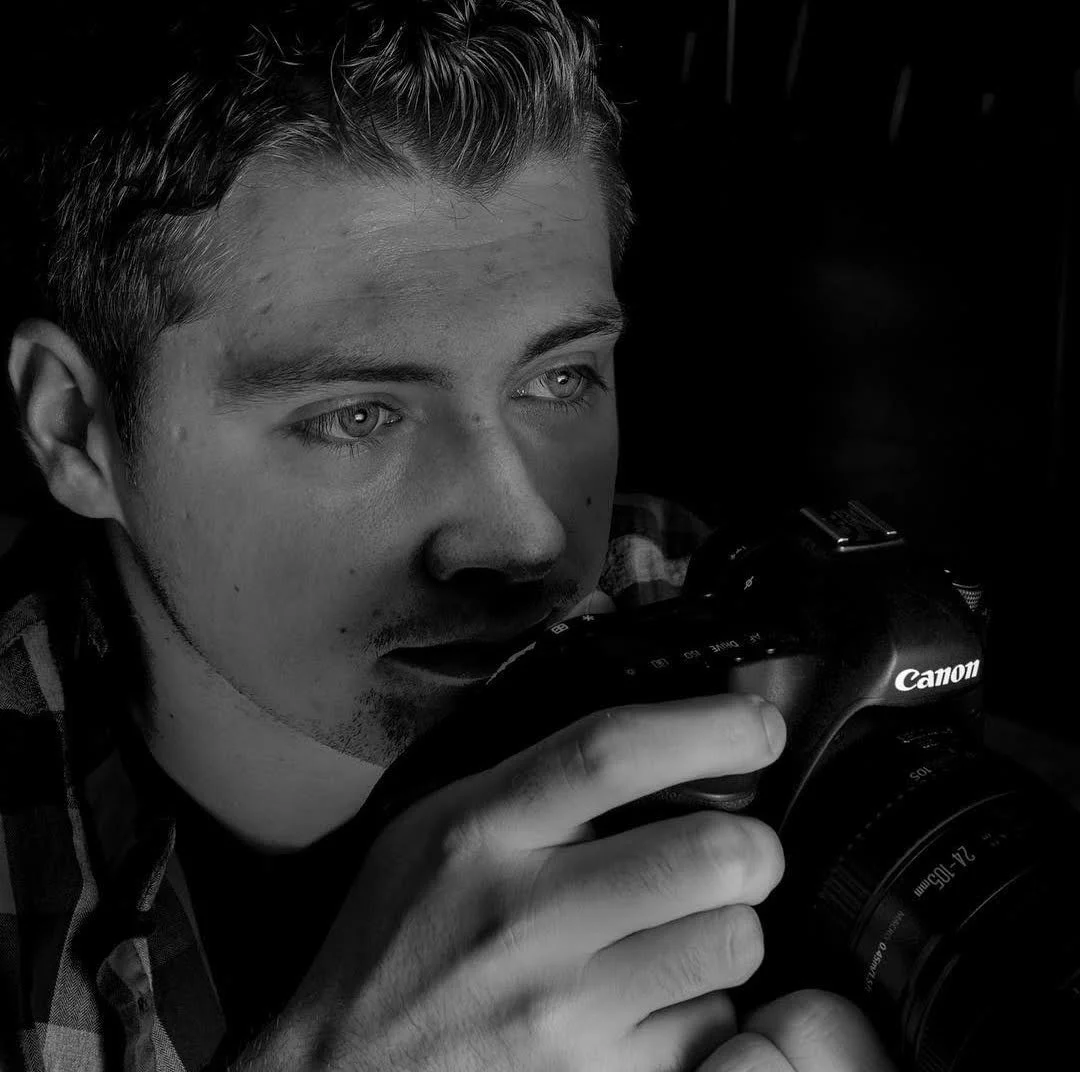 A young man with light-colored eyes holding a Canon camera close to his face, appearing focused, in a black-and-white photograph.