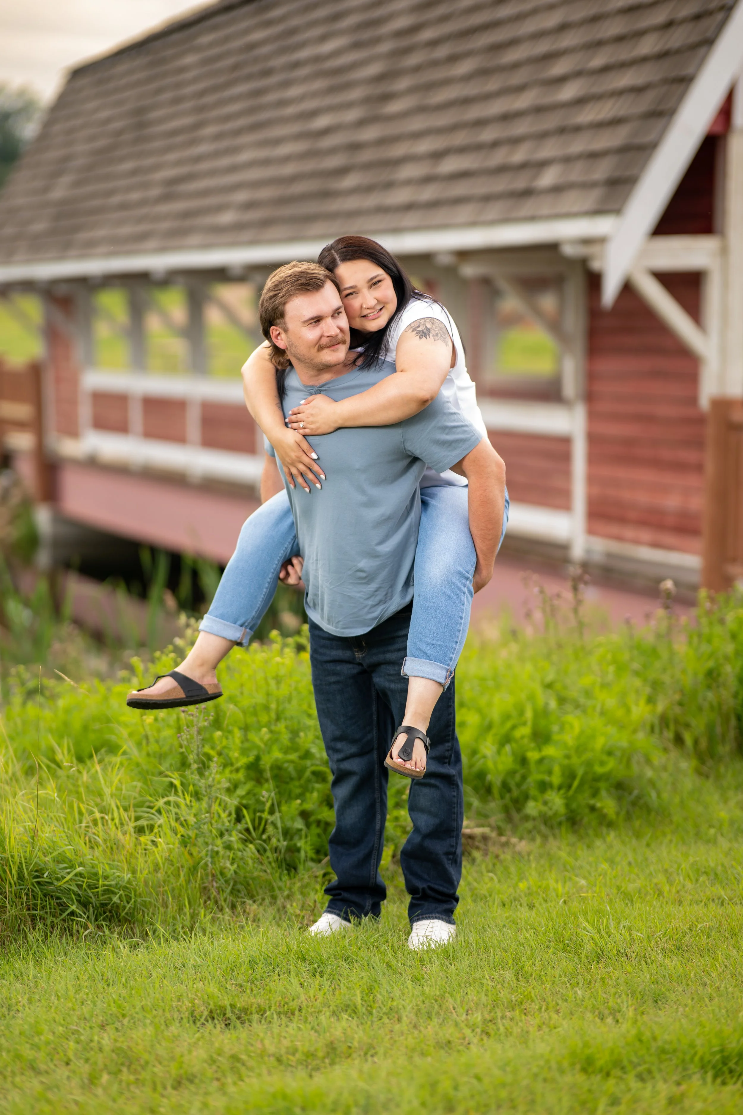 A couple is outdoors, with the man giving the woman a piggyback ride. They are smiling, and there is a wooden house in the background. The woman has dark hair, a tattoo on her arm, and is wearing a white shirt and jeans. The man has light brown hair,