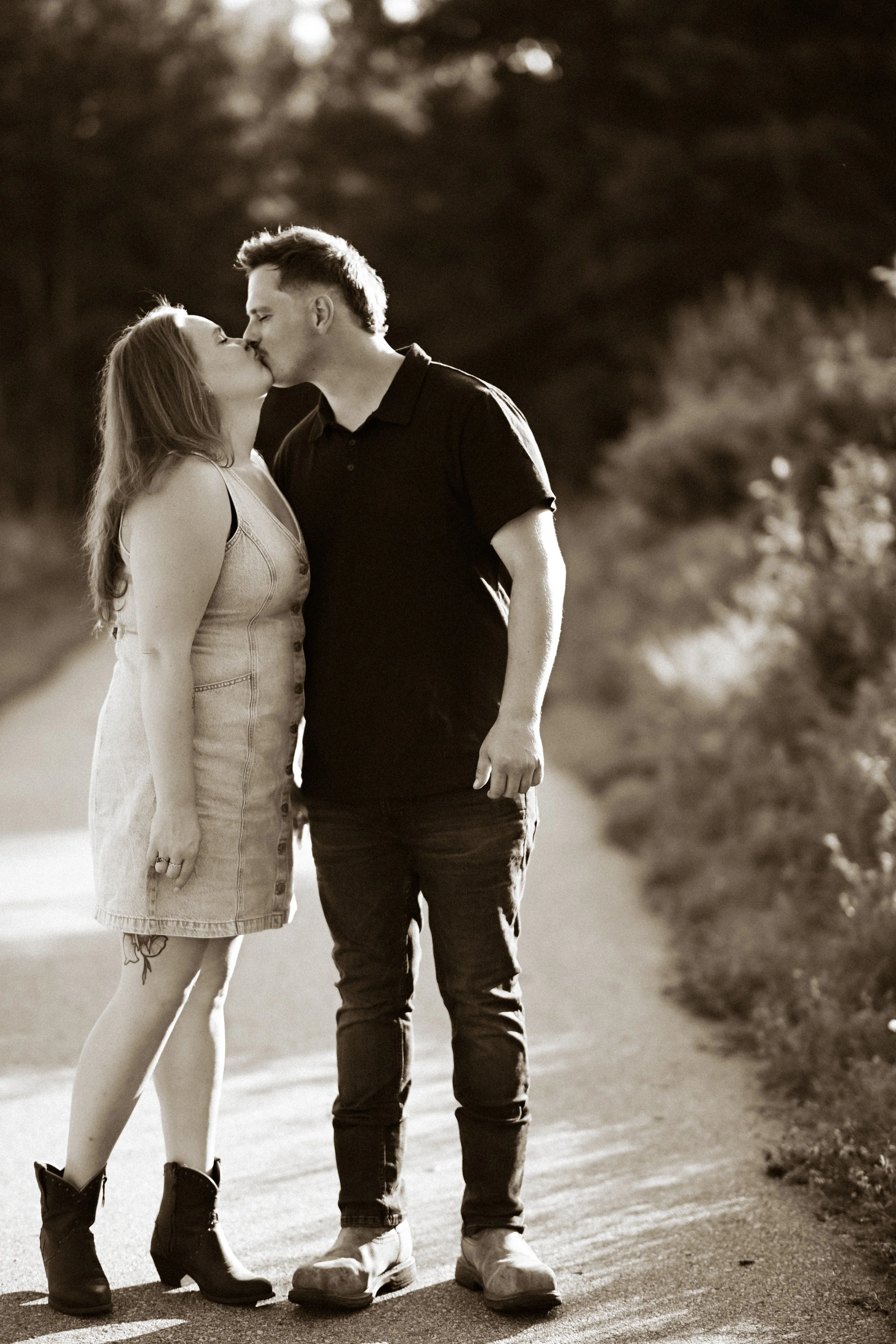 A black and white photo of a man and woman kissing outdoors on a sunny day. The woman has long hair and is wearing a sleeveless dress and boots, while the man is dressed in a polo shirt and jeans. They are standing on a dirt path with trees in the ba