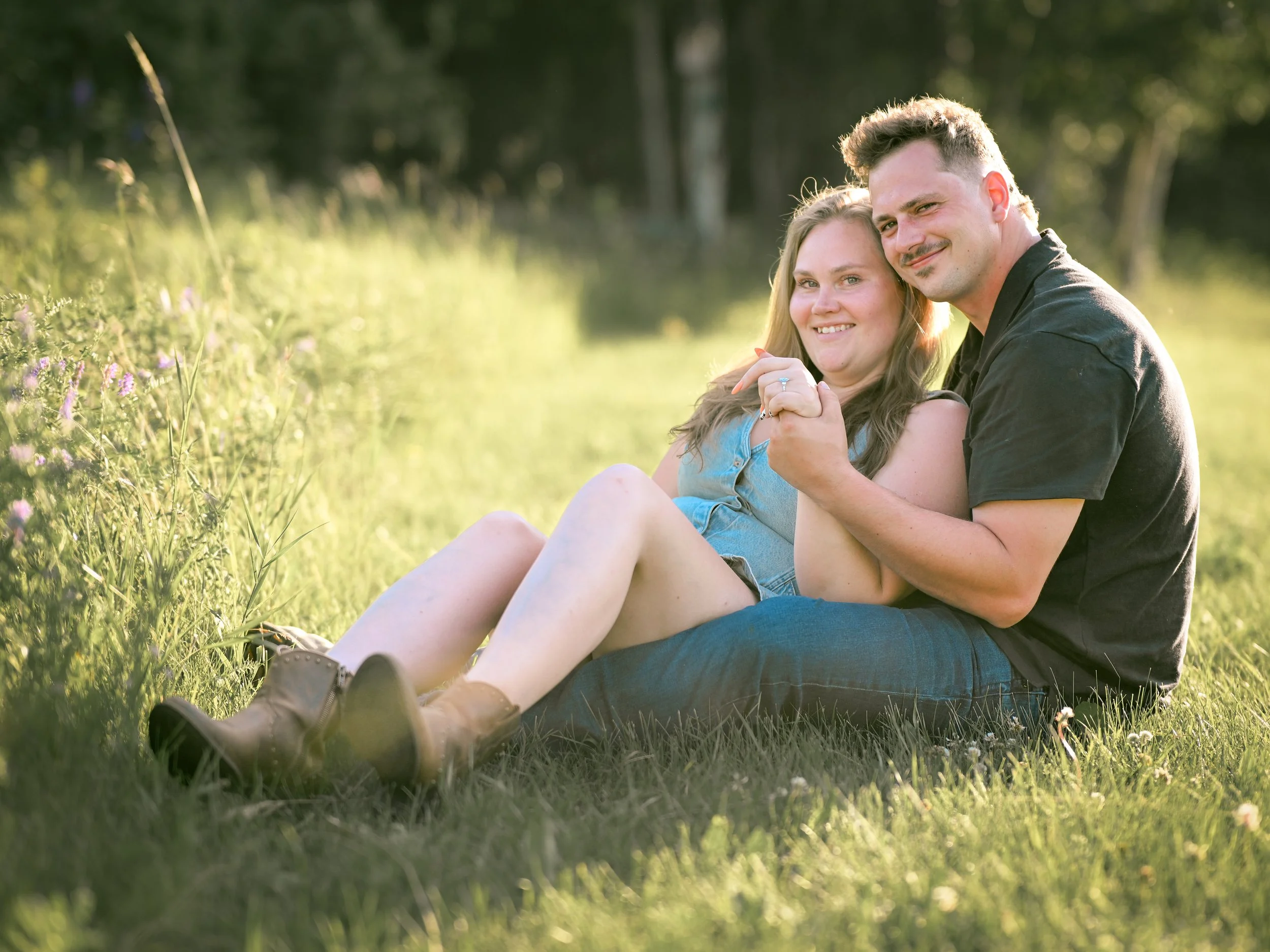 A couple sitting on the grass in a park, smiling and enjoying each other's company, with trees and sunlight in the background.
