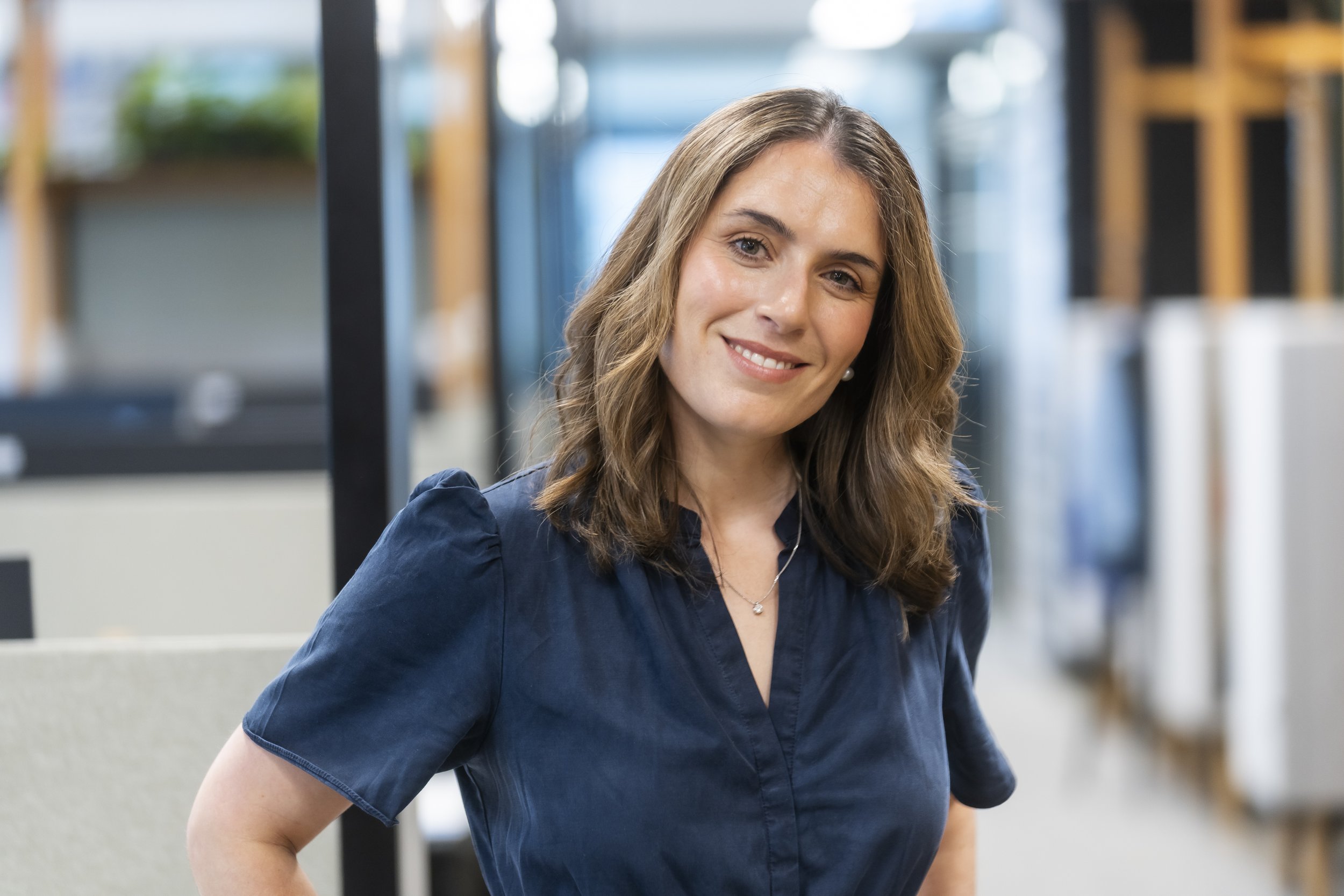 A woman with long, wavy brown hair and wearing a navy blue shirt smiling and looking at the camera in an indoor setting with blurred background.