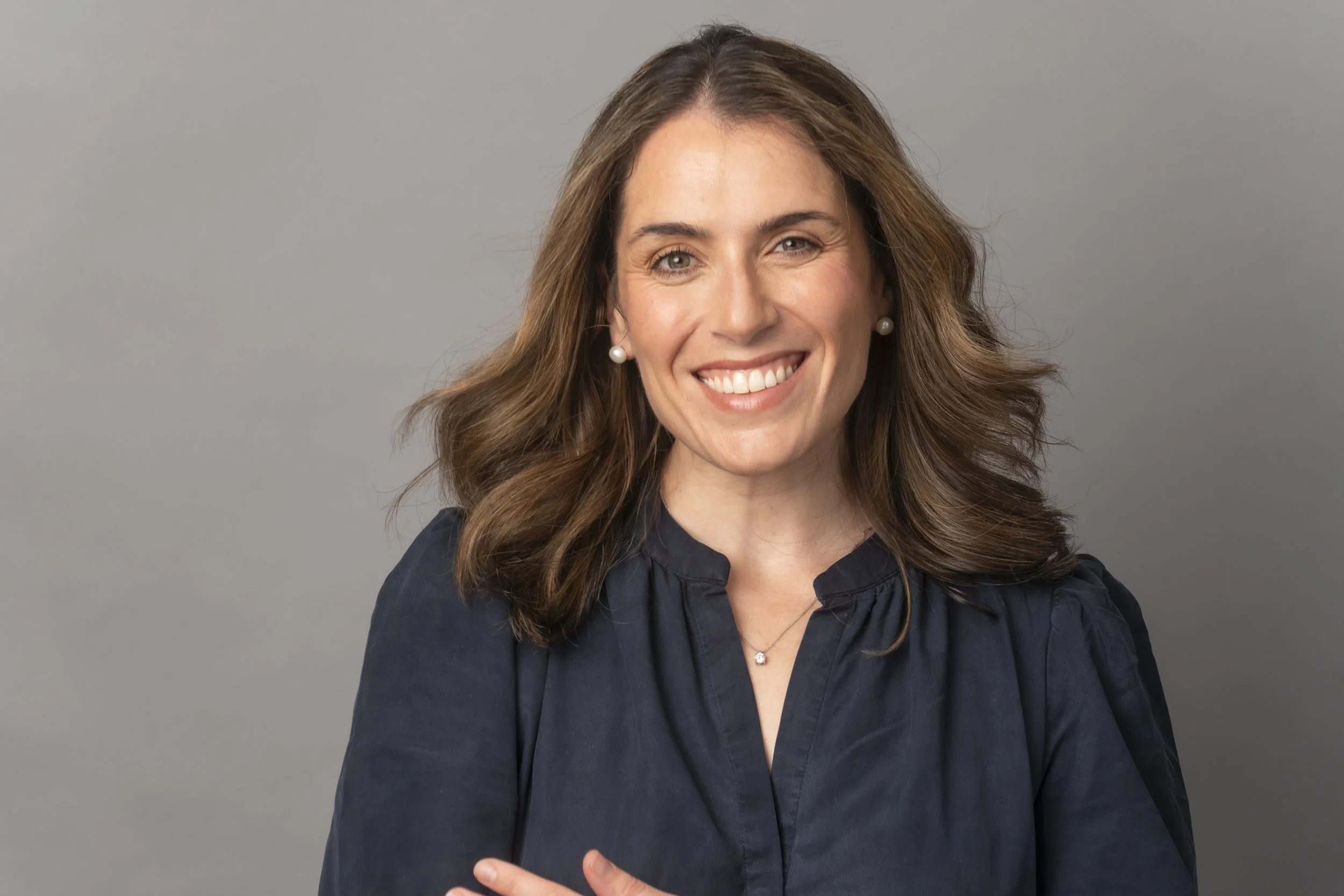 A smiling woman with long wavy brown hair wearing pearl earrings and a dark navy blouse against a light gray background.