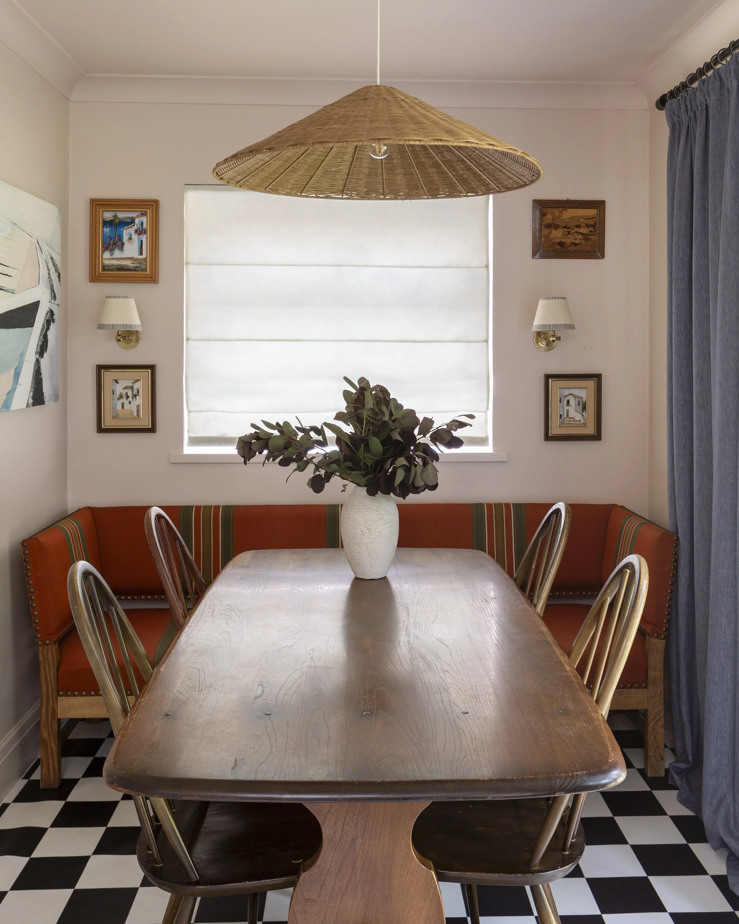 A wooden dining table with a white vase filled with green leaves, surrounded by wooden chairs inside a room with cream-colored walls, framed artwork, wall-mounted lamps, a large window with blinds, and blue curtains on the right.