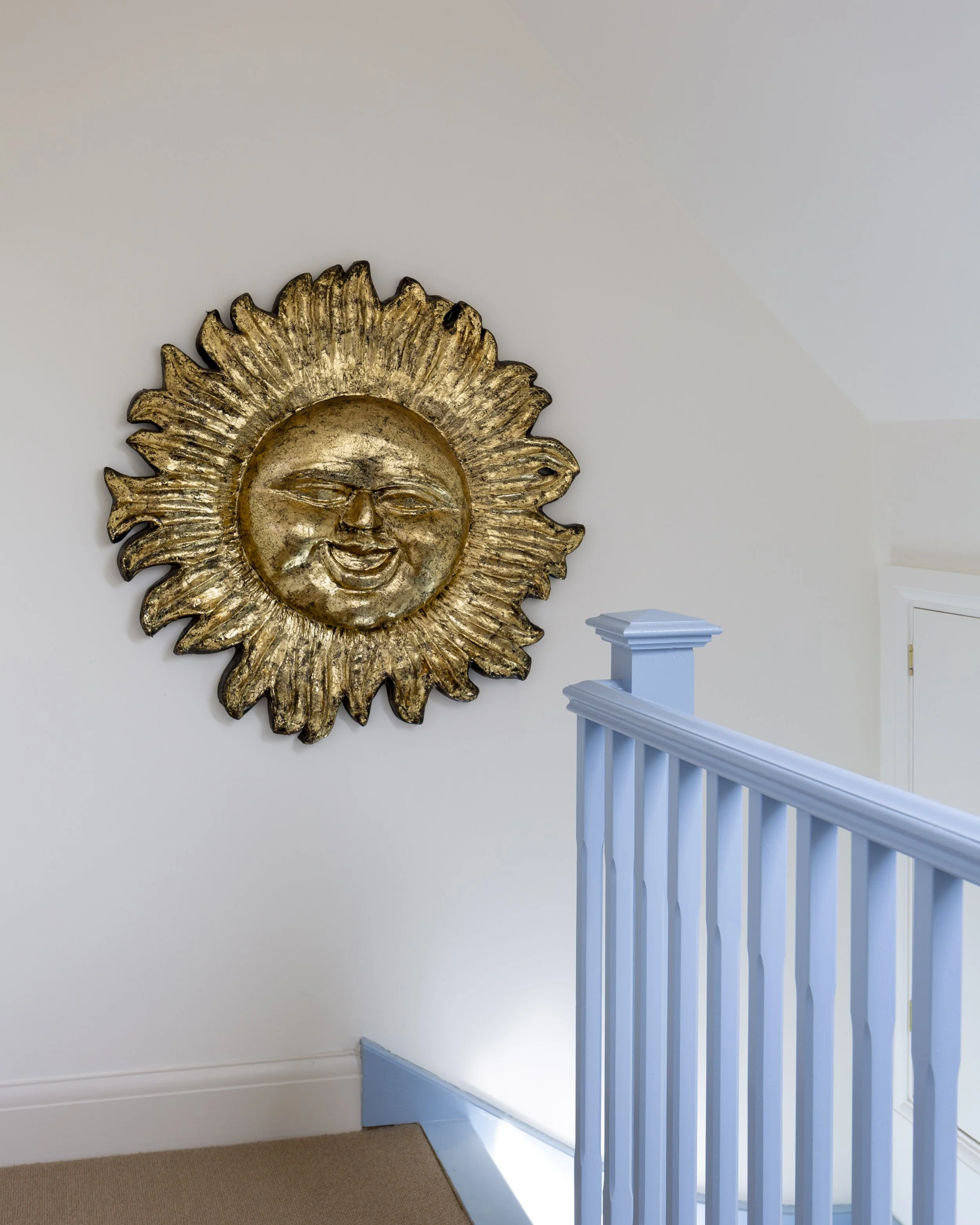 A gold sun sculpture with a smiling face hanging on a white wall beside a white staircase banister.