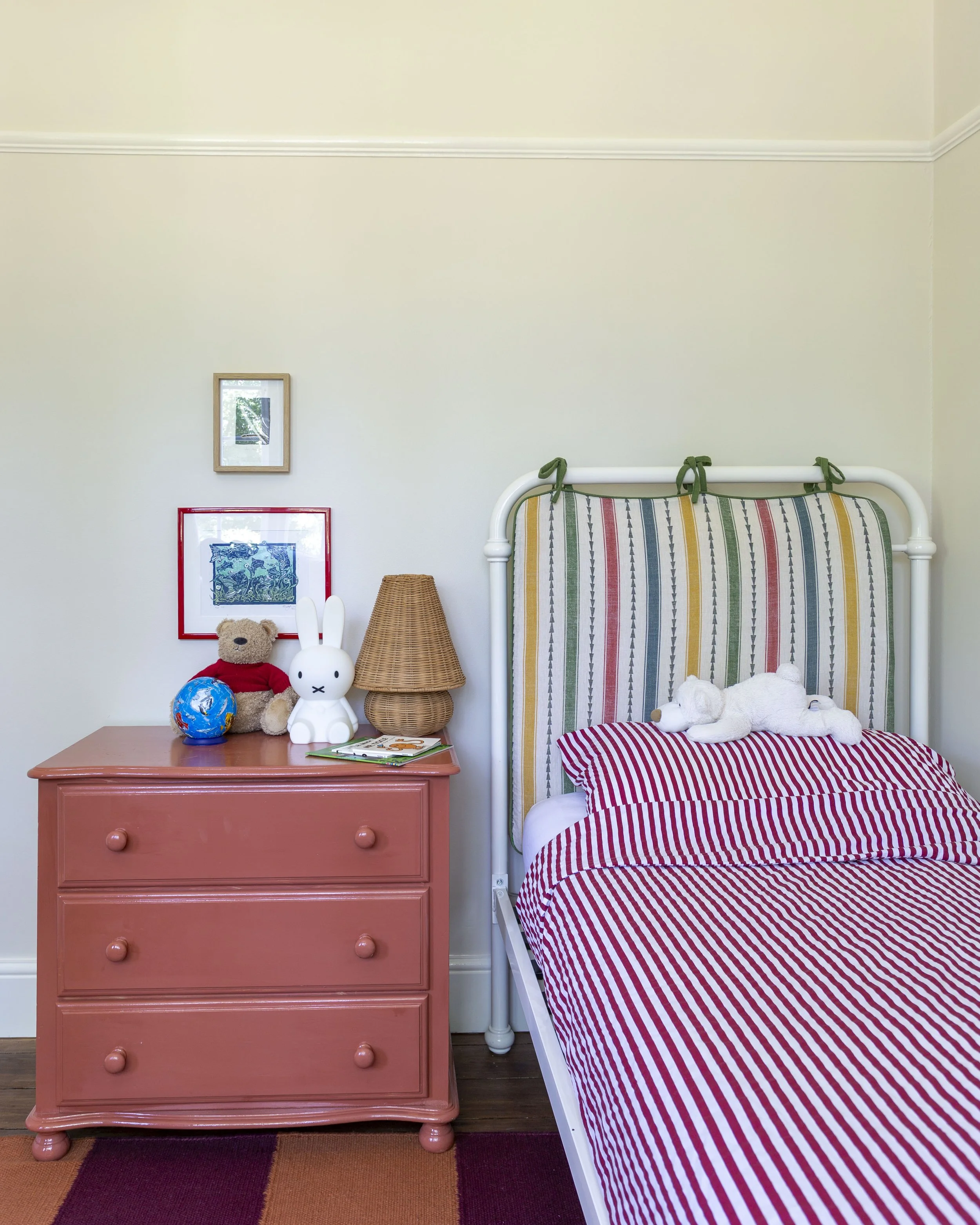 children's room with off white walls, a red chest of drawers. iron bed with stripy headboard