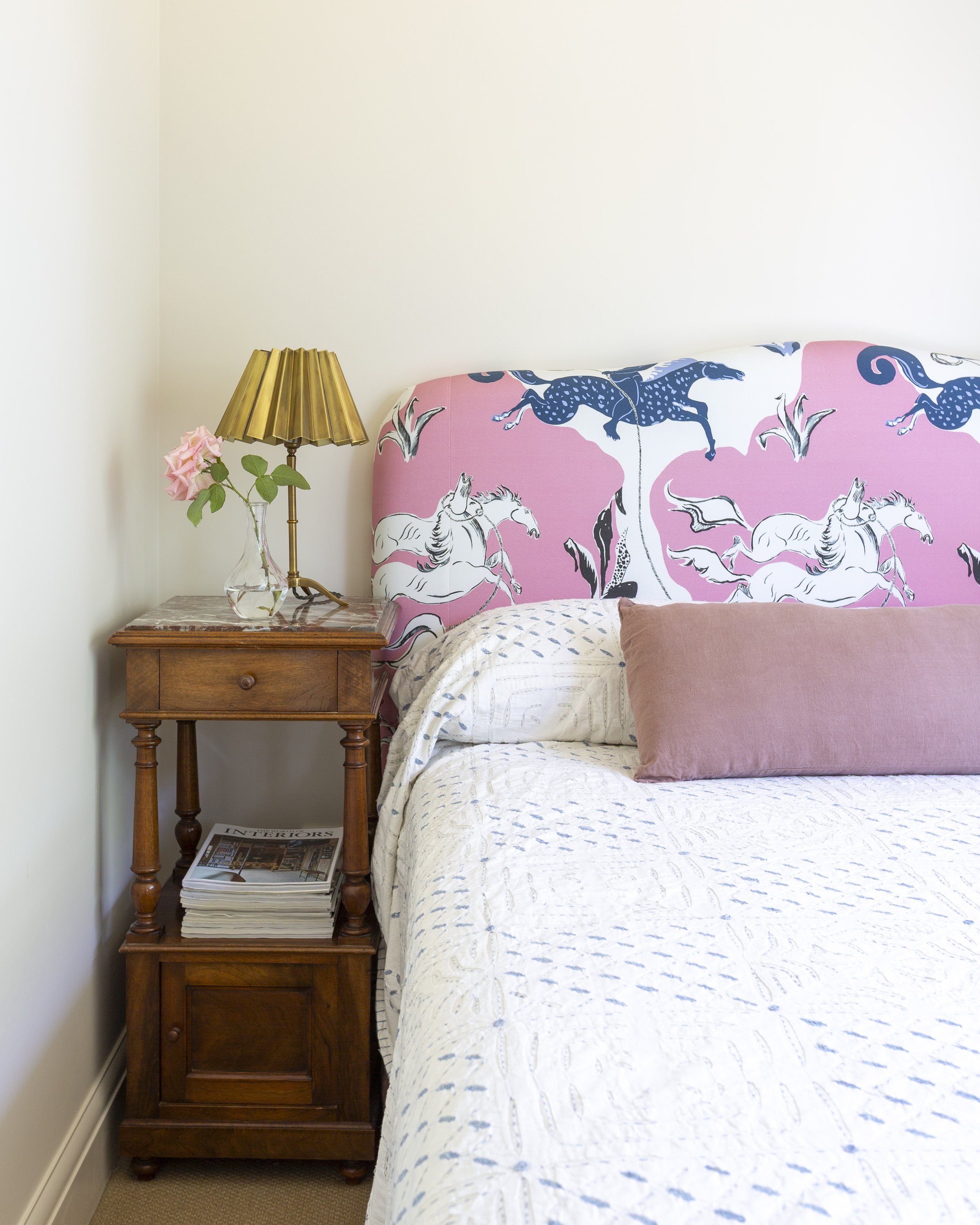 A bedroom corner with a wooden nightstand holding a glass vase with pink roses and a vintage gold-colored lamp. The bed features a pink and white headboard with a horse and floral pattern, and is covered with a white quilt and a pink pillow.