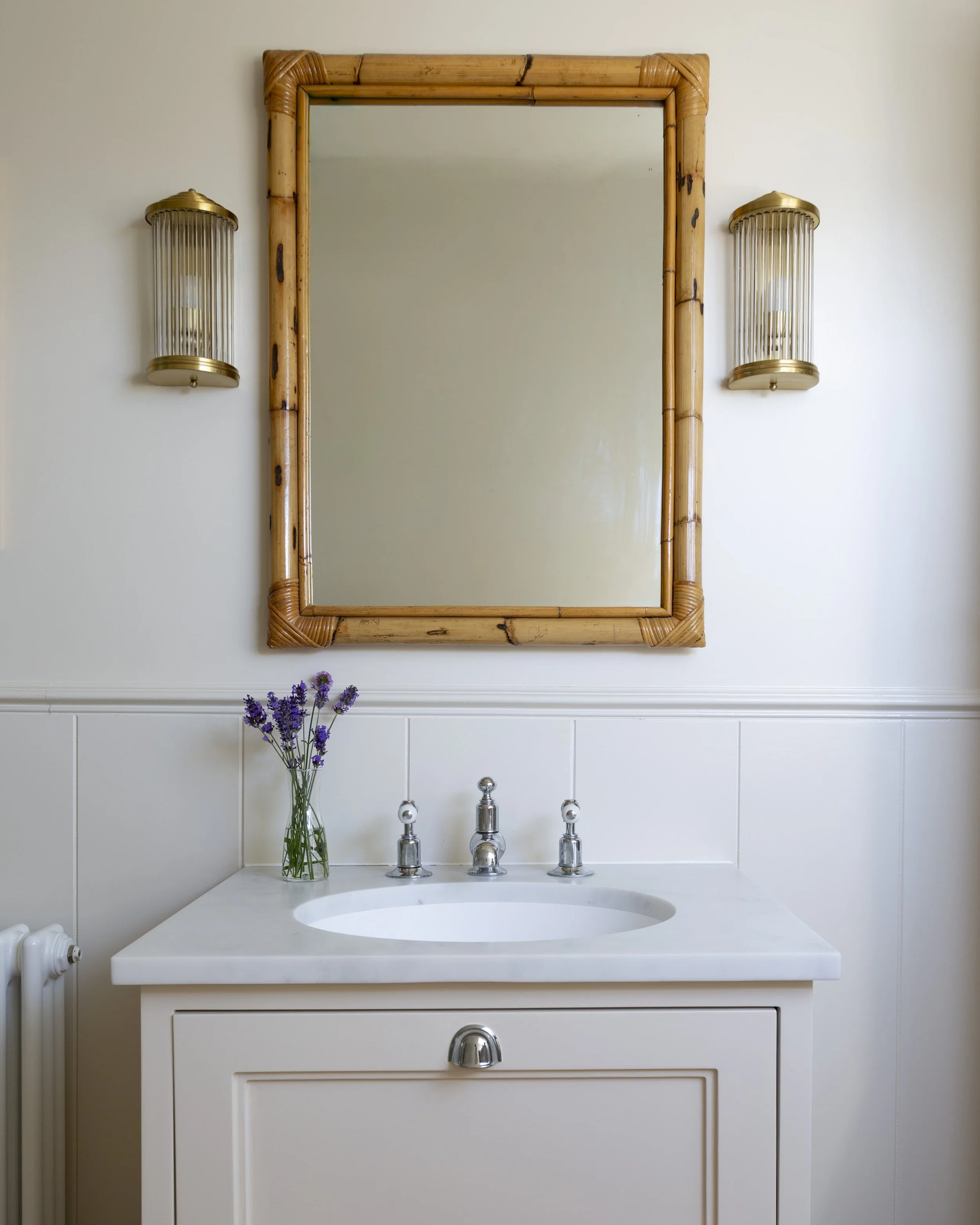 A bathroom vanity with a white countertop, a built-in sink, and a cabinet with a silver knob. A tall, clear glass vase with purple flowers is placed on the counter. Above, a large mirror with a bamboo frame is flanked by two gold wall-mounted light fixtures with vertical bars.