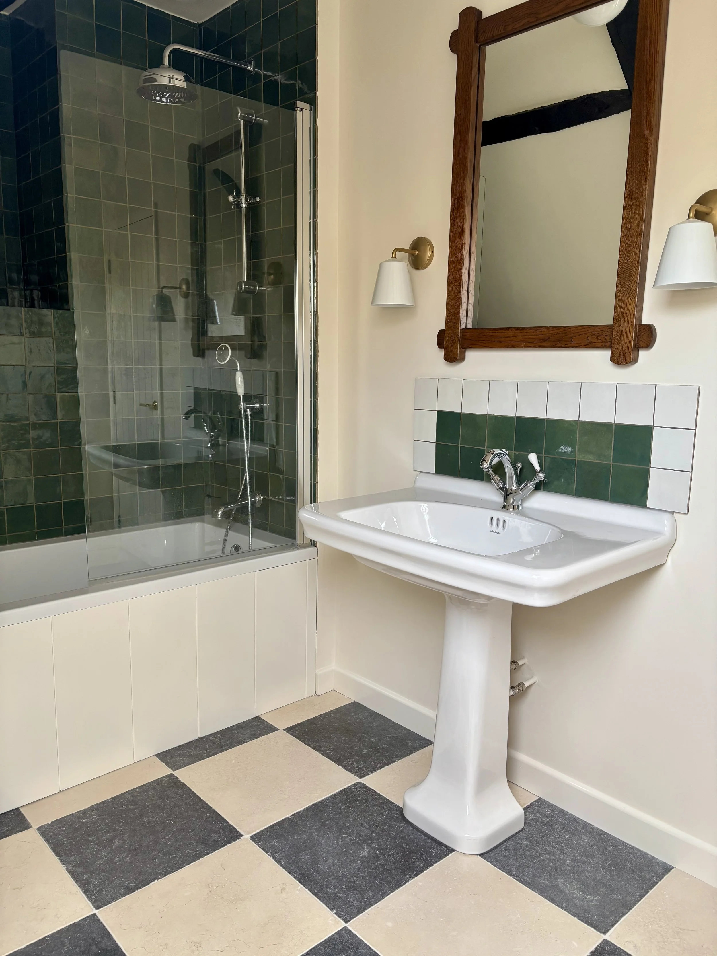 bathroom with limestone chequerboard floors, a sink with a green and white cplashback.  Wall lights with vintage brutalist mirror. bath with green zellige tiles