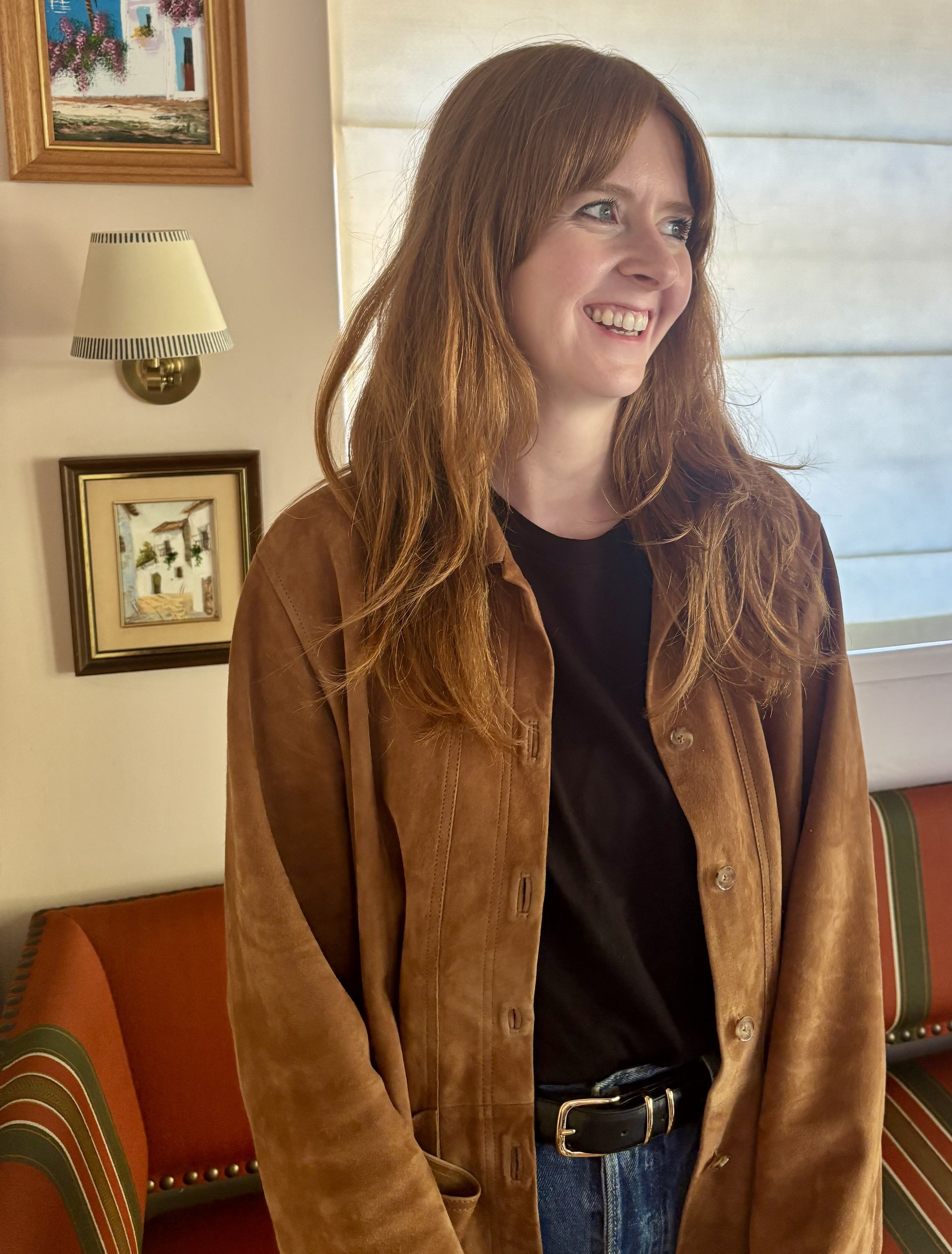 A woman with long red hair smiling, wearing a brown jacket over a black shirt, standing indoors near a wall with paintings and a window with blinds.