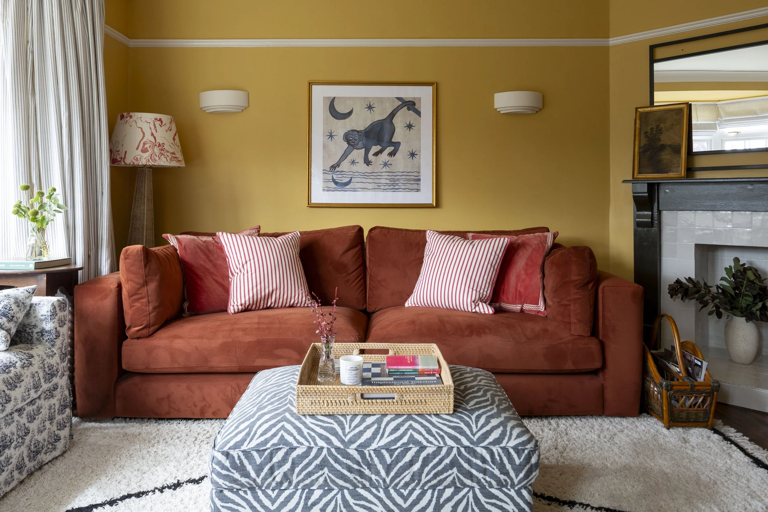 Living room with a rust-colored sofa with red and white striped pillows, a patterned ottoman, a wooden side table with plants and books, a tall floor lamp with a red and white patterned shade, yellow walls with framed wall art, a fireplace with a black mantle and mirror above, and decorative items like a small painting and plants.