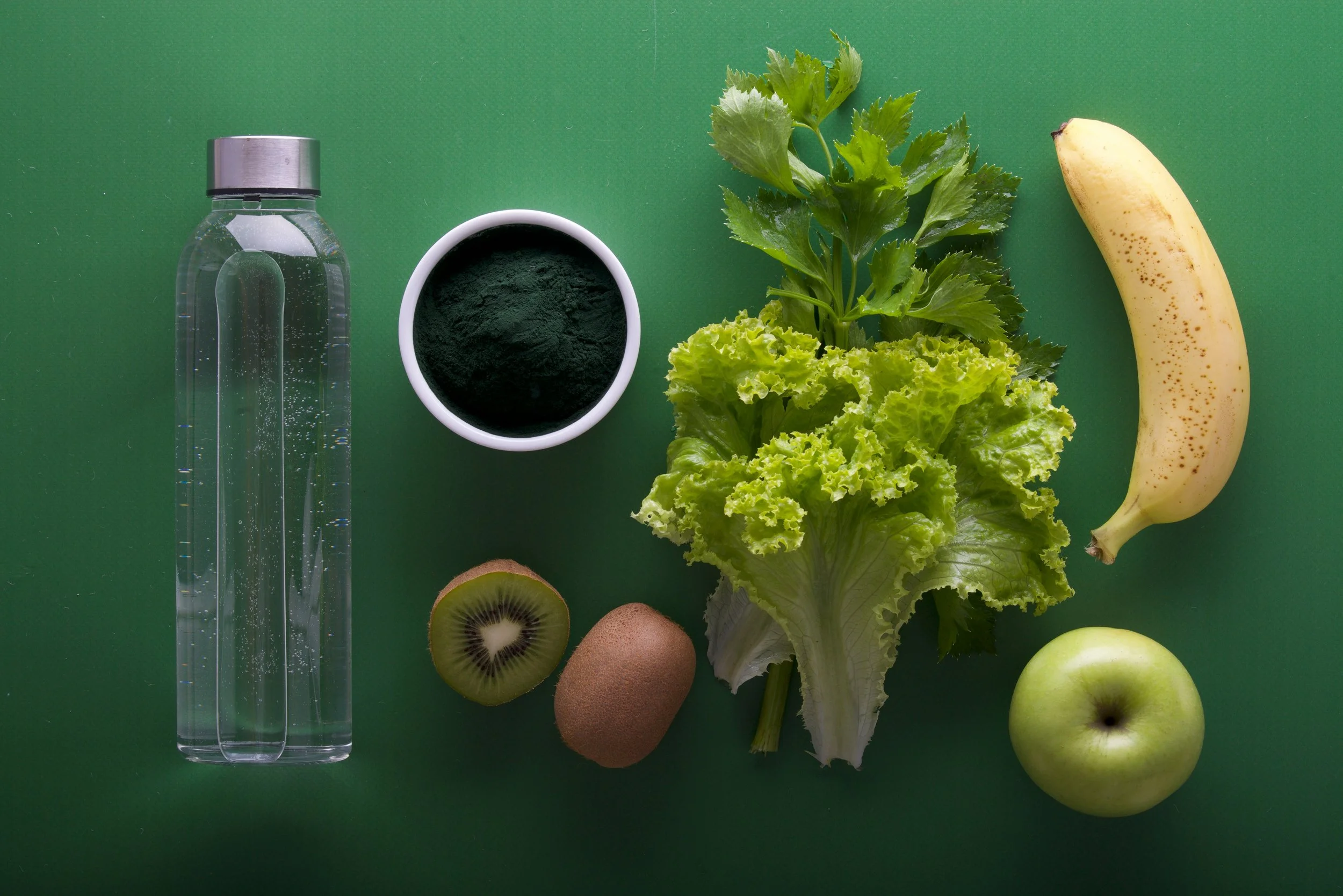 A green background with a clear water bottle, a white cup of spirulina powder, fresh leafy greens, a banana, a kiwi, and green apples.