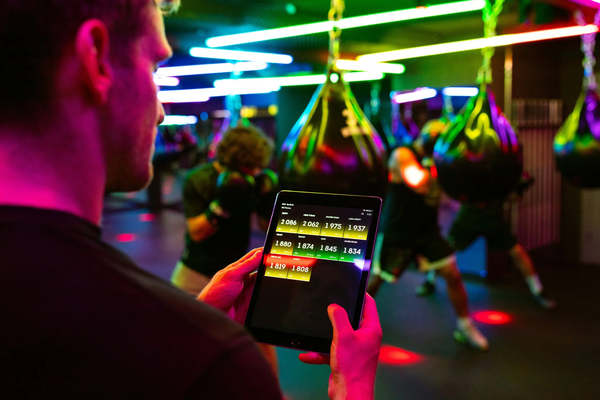 A person holding a smartphone in a dim, colorful arcade or amusement park with neon lights. The screen displays scoreboard results for a game, with names and scores. In the background, there is a person wearing headphones standing near a carousel ride with neon lights.