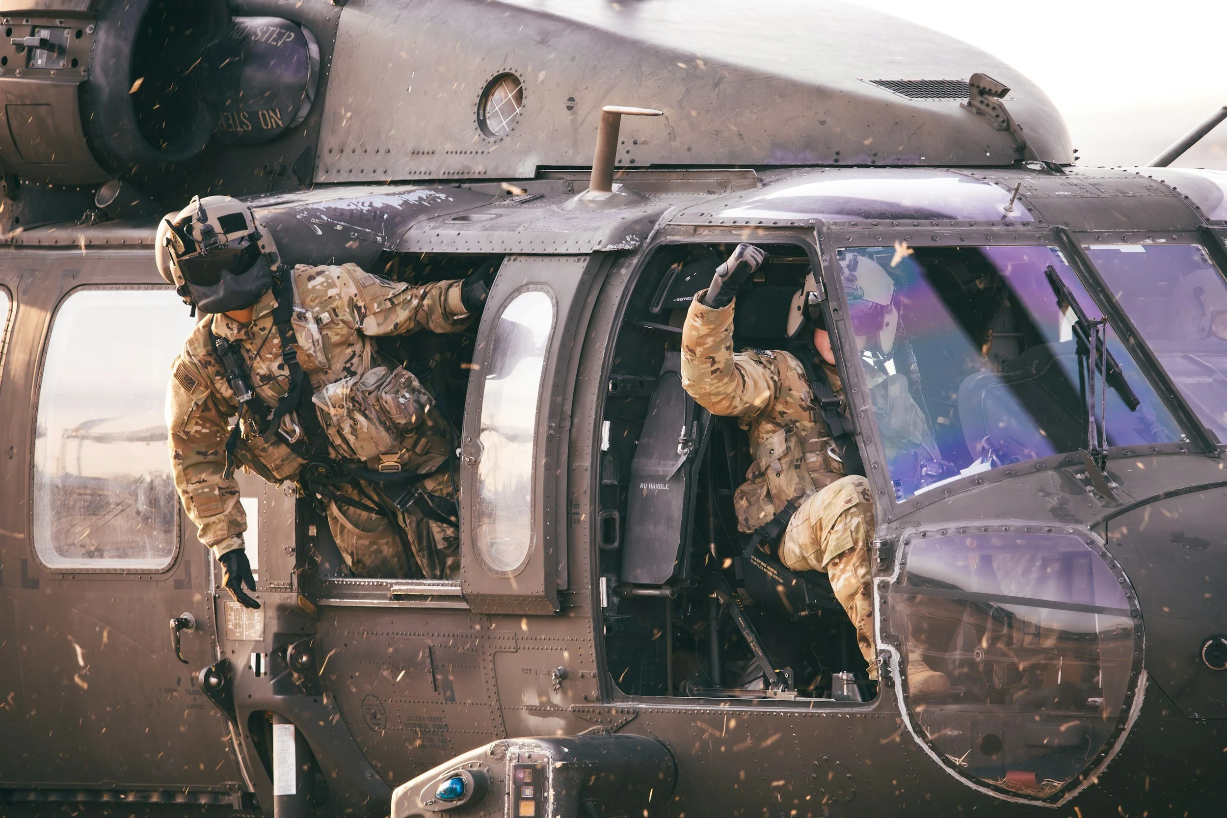Two soldiers in camouflage uniforms and helmets sitting inside a military helicopter. The helicopter is black with various panels, rivets, and a large window, with some sparks or debris flying around.