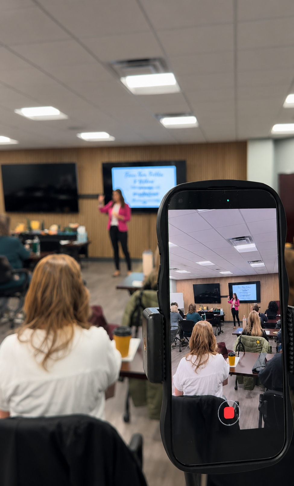 A woman giving a presentation in a conference room, seen on a screen through a smartphone's recording app, with audience members seated and listening.